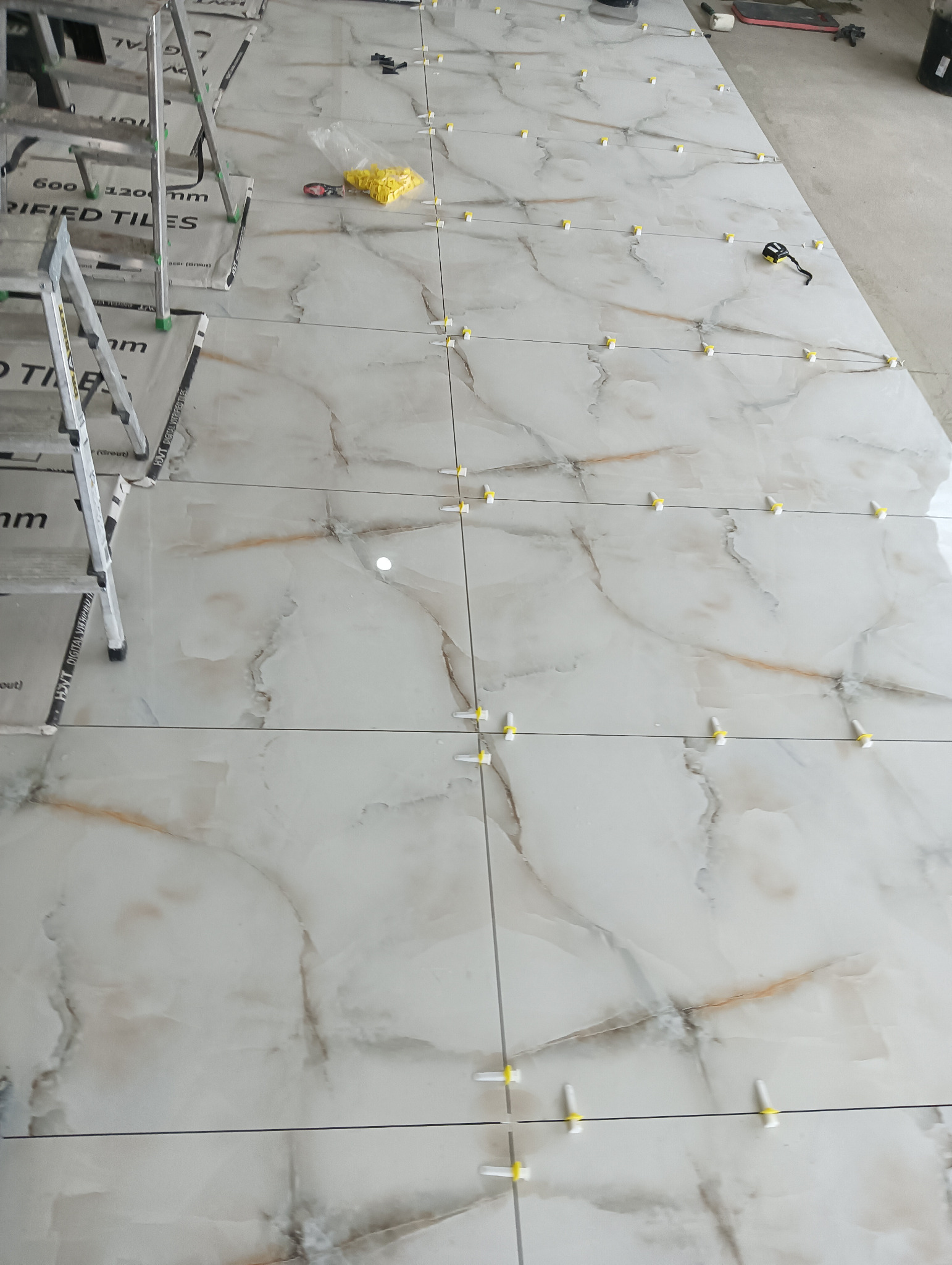 Układanie płytek podłogowych imitujących marmur, z użyciem poziomujących klipsów i klinów, widoczne narzędzia i drabina w tle.