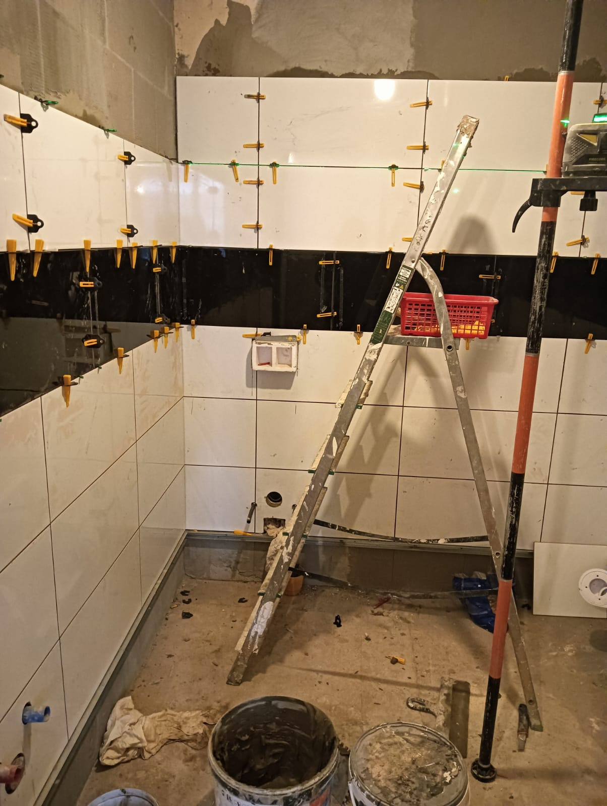 Wnętrze łazienki w trakcie układania białych i czarnych płytek na ścianach, widoczna drabina, poziomica laserowa i wiadra z zaprawą.