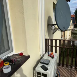 Montaż klimatyzatora na balkonie, widoczne narzędzia, skrzynka złączek i telefon na obudowie jednostki zewnętrznej, obok okna i anteny satelitarnej.