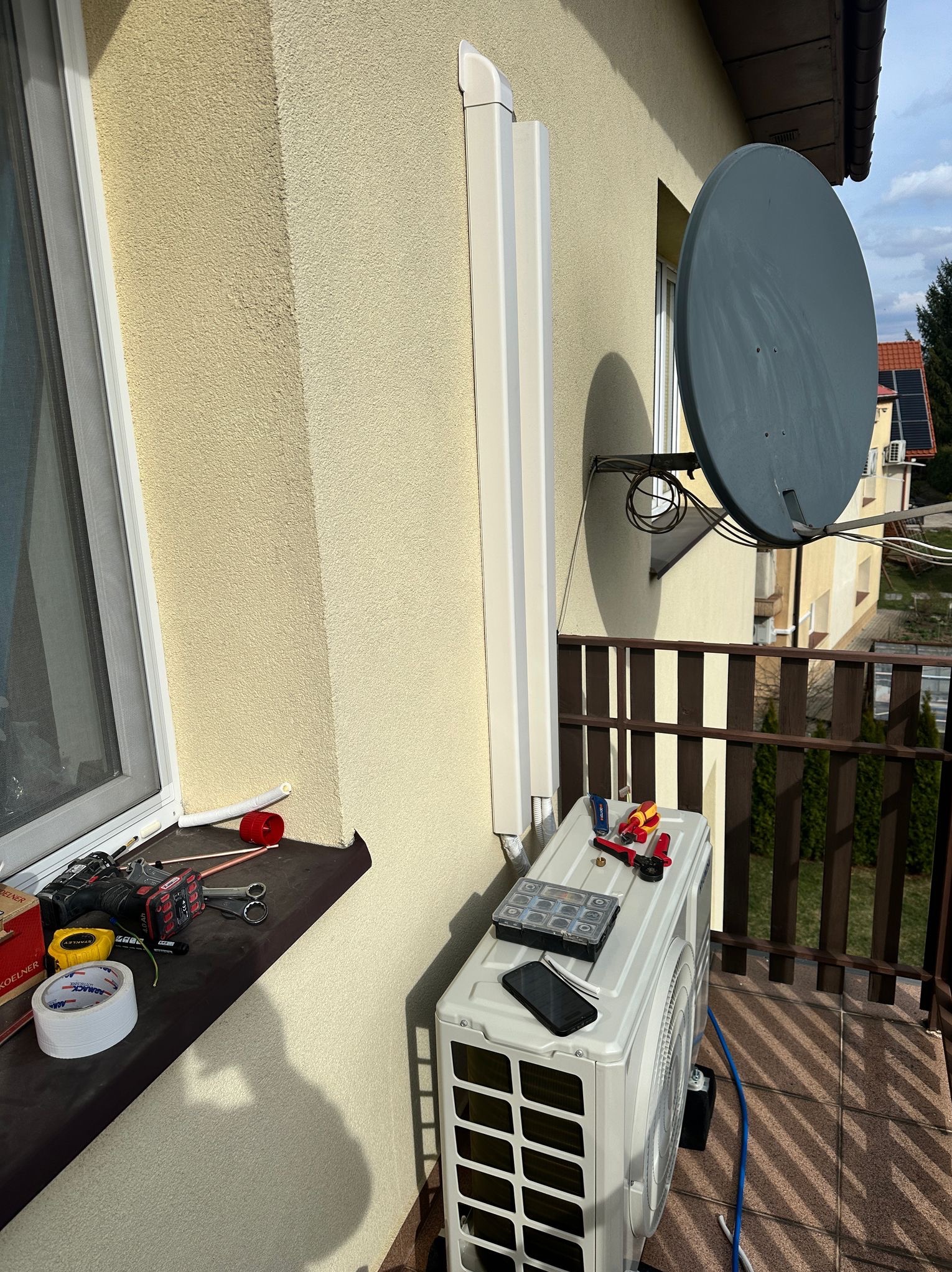 Montaż klimatyzatora na balkonie, widoczne narzędzia, skrzynka złączek i telefon na obudowie jednostki zewnętrznej, obok okna i anteny satelitarnej.