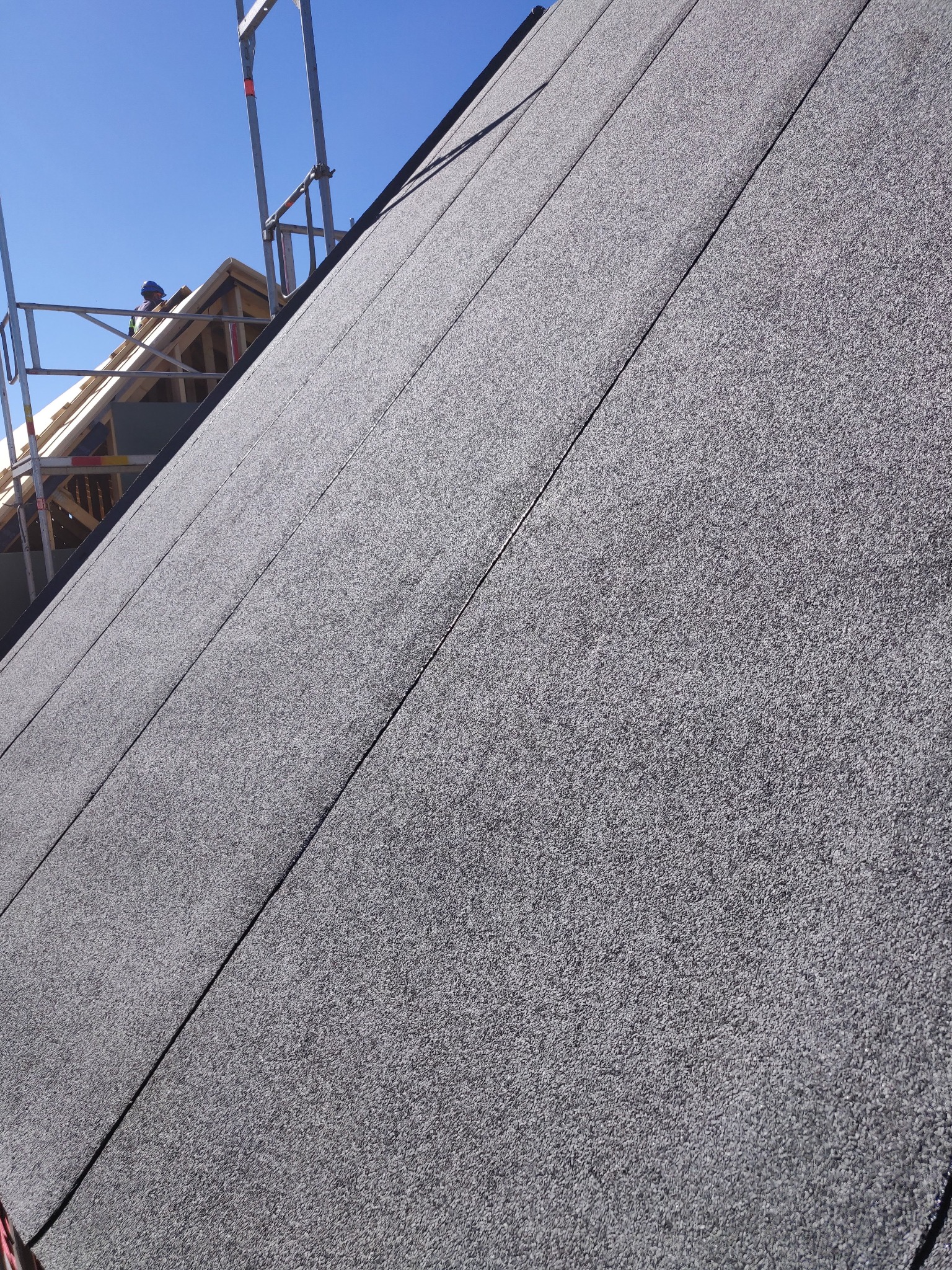 Szary dach pokryty papą termozgrzewalną, widoczny fragment rusztowania i pracownik w oddali, błękitne niebo w tle. Układanie papy na dachu.