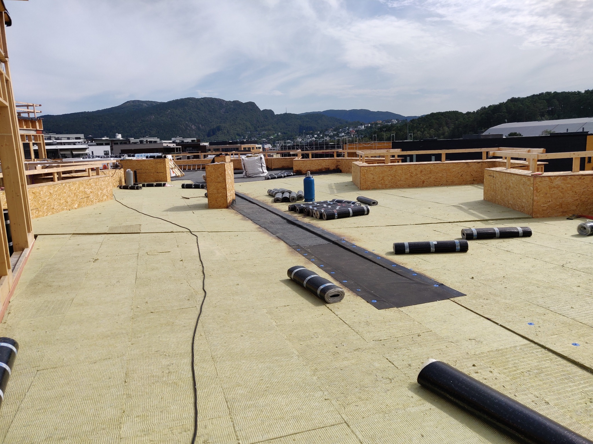 Izolacja termiczna dachu płaskiego z rolkami papy na tle górskim. Widoczne drewniane elementy konstrukcyjne i częściowo ułożona warstwa izolacyjna.