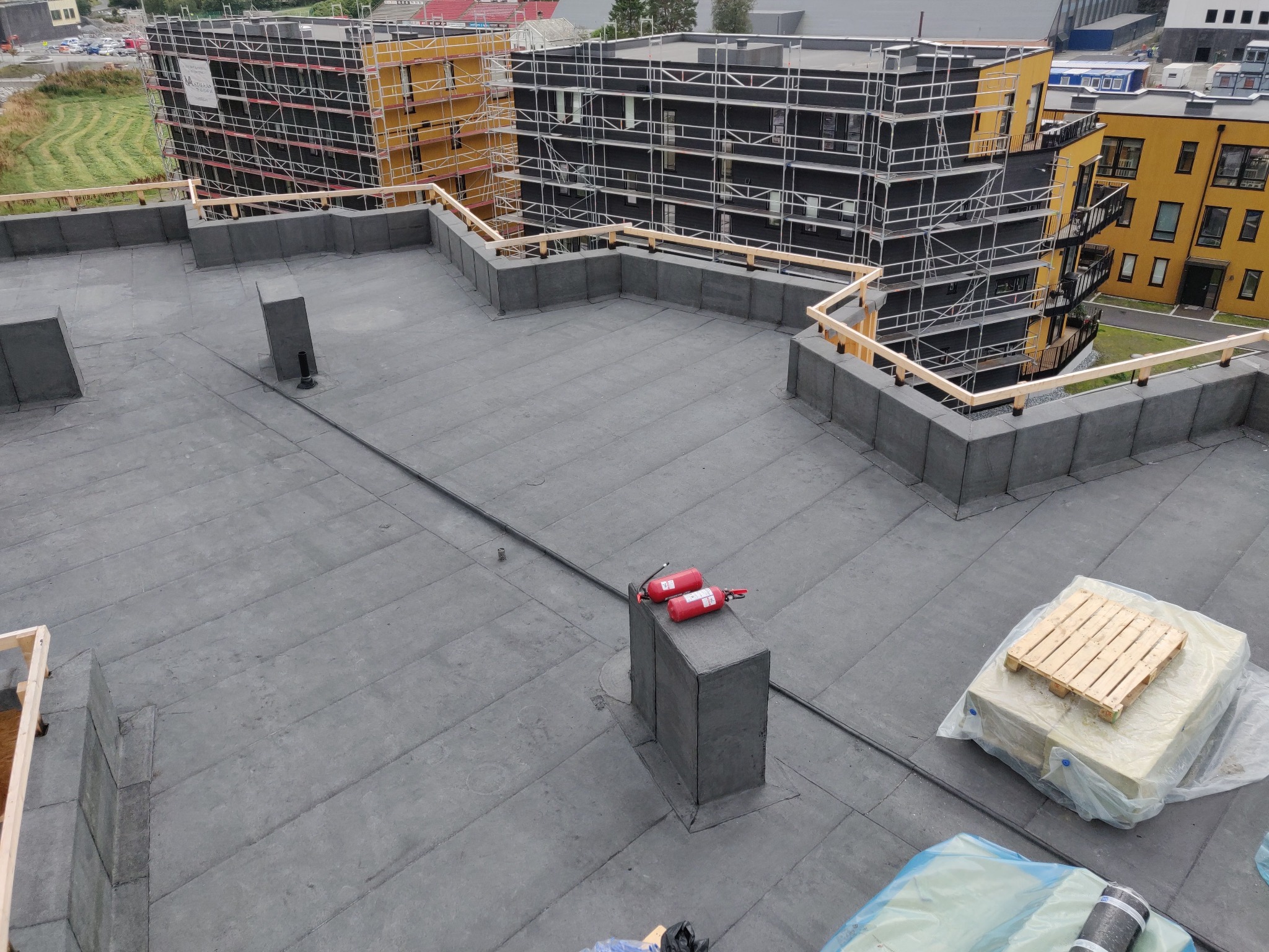 Wykończony, płaski dach budynku wielorodzinnego z zabezpieczeniami, widoczne materiały izolacyjne i gaśnice. W tle budynki w rusztowaniach.