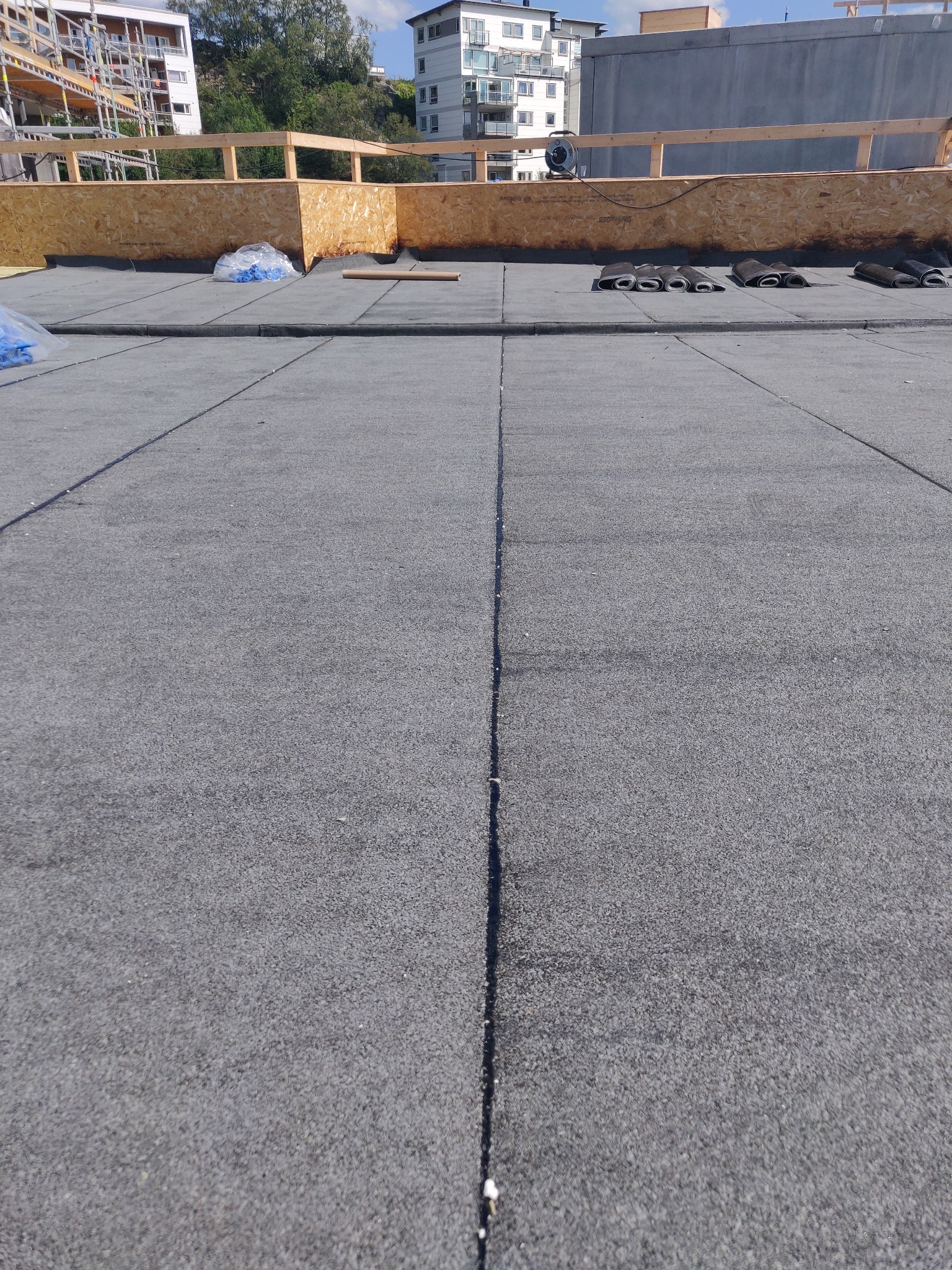 Szary, płaski dach pokryty papą, z widocznymi łączeniami. W tle drewniana konstrukcja zabezpieczająca i budynki mieszkalne w oddali. Częściowo zwinięta papa na dachu.