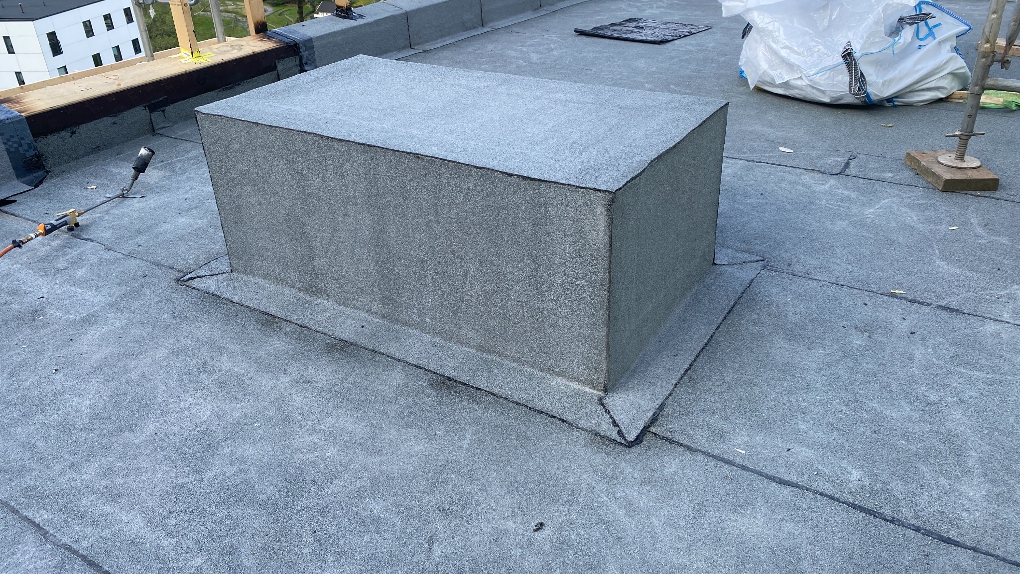 Szczegółowe ujęcie płaskiego dachu pokrytego papą, z wykończonym kominem wentylacyjnym. Widoczne narzędzia dekarskie i worki z materiałami budowlanymi w tle.