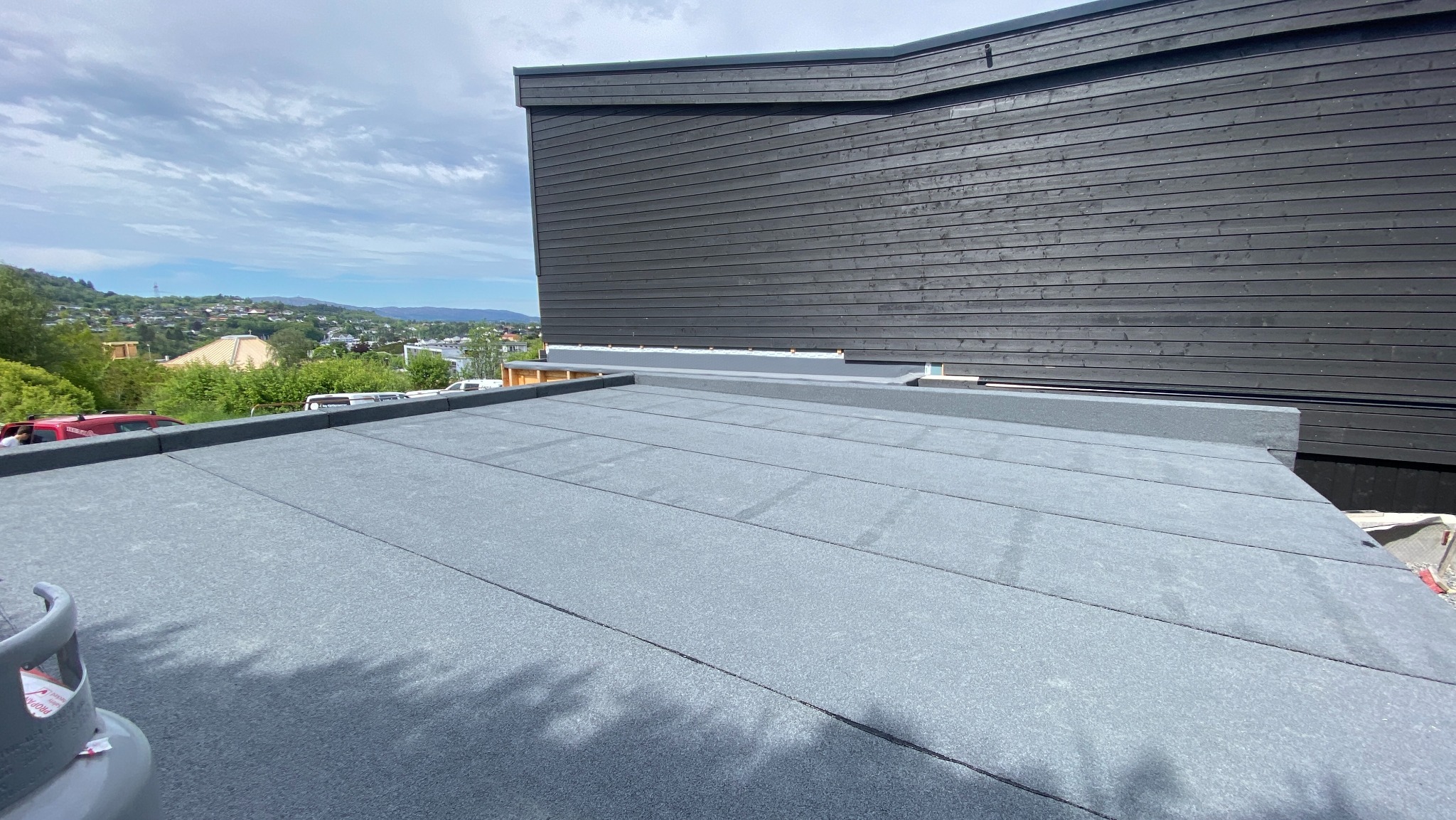 Szary, płaski dach pokryty papą termozgrzewalną na tle ciemnej, drewnianej elewacji i zielonego krajobrazu. Widoczny fragment butli gazowej.