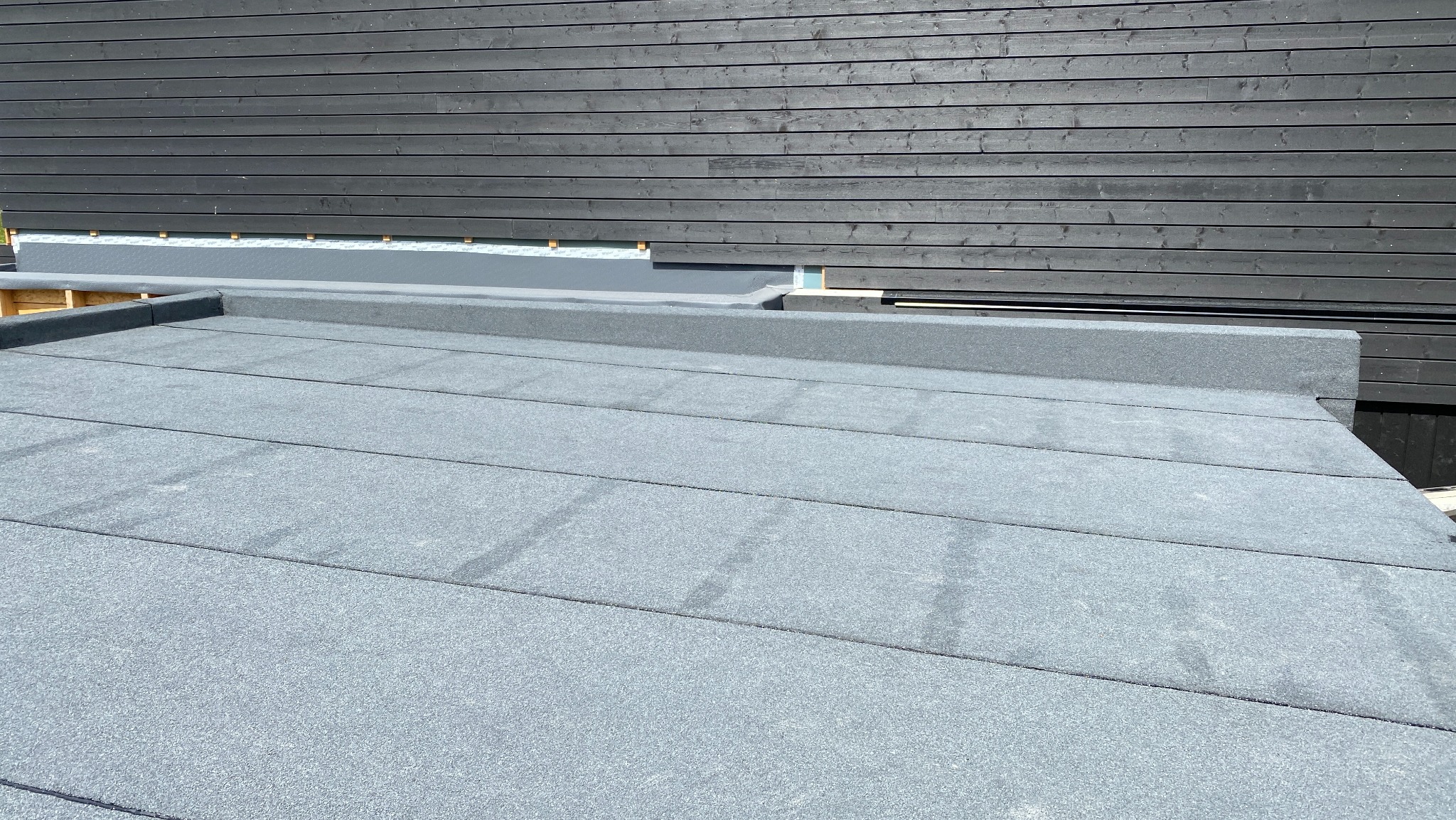 Szary, płaski dach pokryty papą termozgrzewalną, z widocznym fragmentem czarnej, drewnianej elewacji. Detal wykończenia dachu.