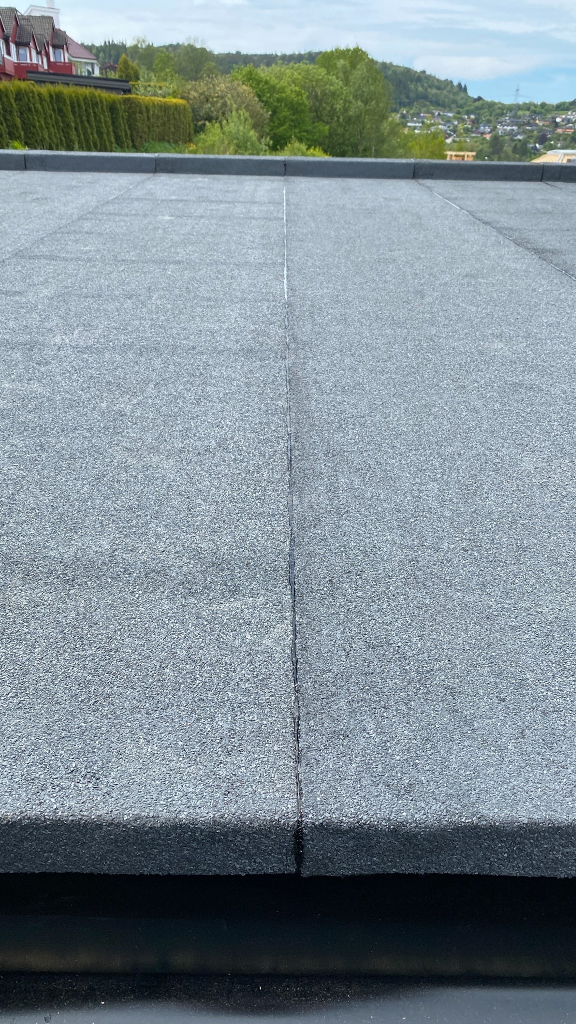 Szczegółowe ujęcie płaskiego dachu pokrytego papą z widocznymi łączeniami, na tle zieleni drzew i zabudowań w oddali, ukazujące precyzję wykonania.