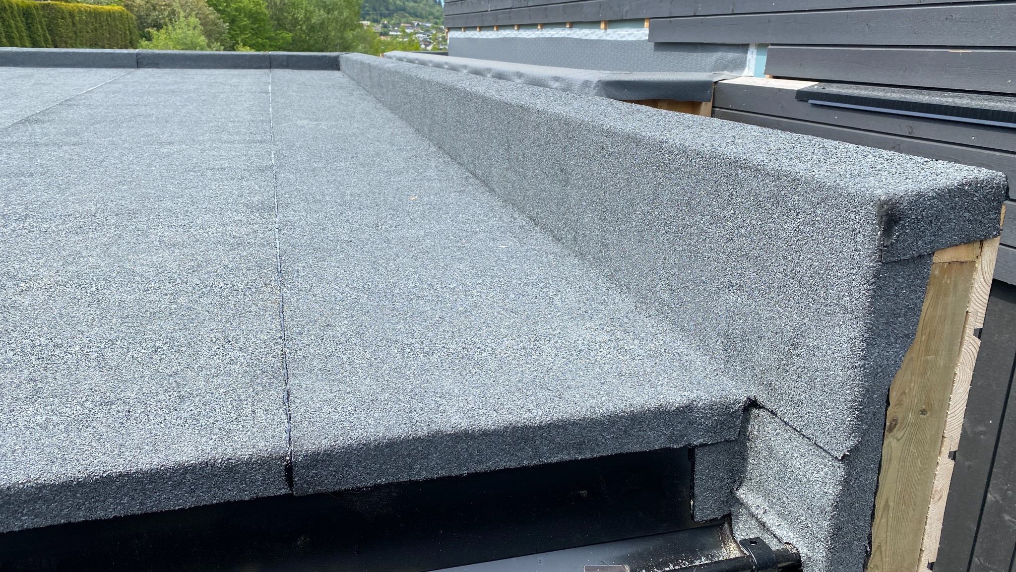 Szczegółowe ujęcie płaskiego dachu pokrytego szarą papą z widocznymi obróbkami blacharskimi i elementami konstrukcyjnymi. Widok z góry na fragment dachu.