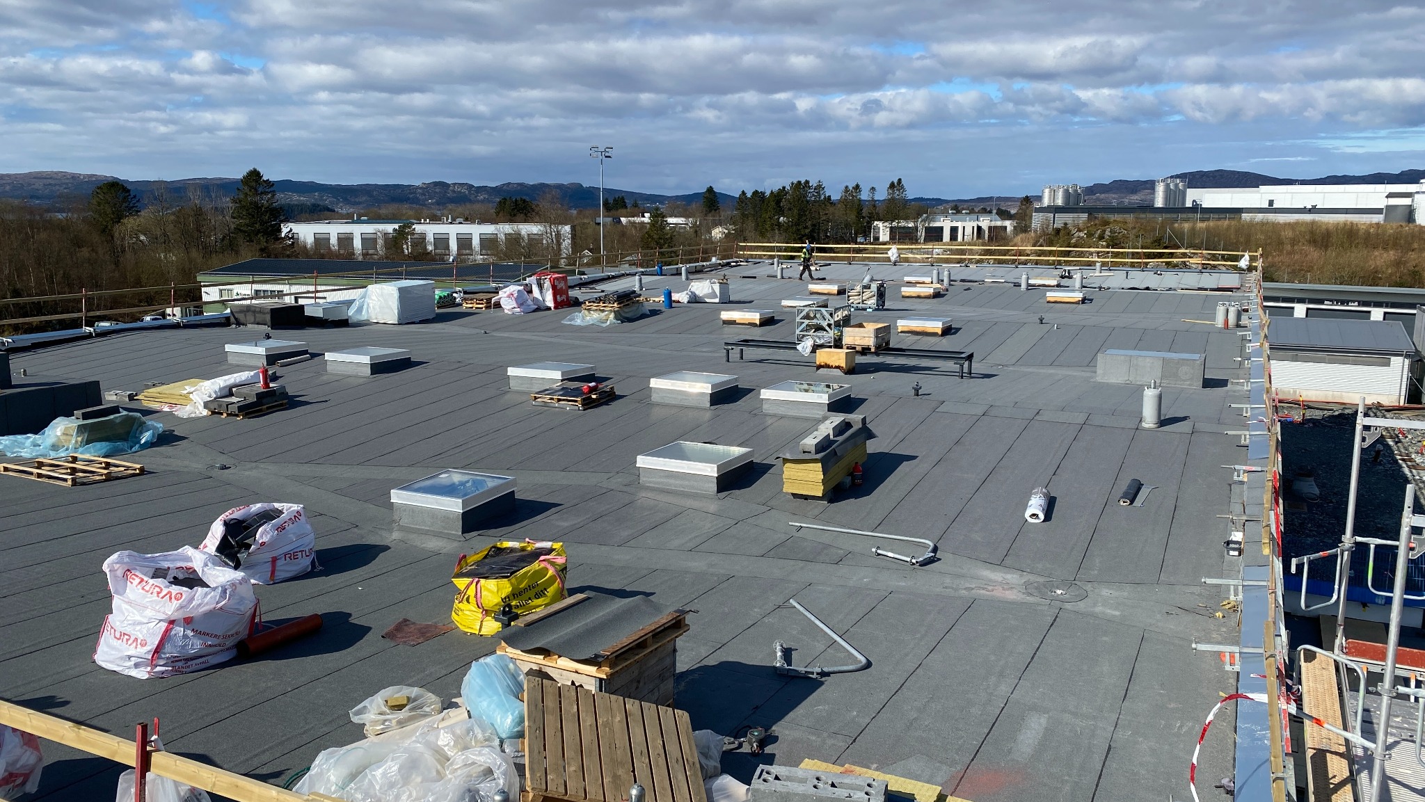 Rozległy, płaski dach pokryty szarą membraną, z licznymi świetlikami i materiałami budowlanymi. W tle budynki i pagórkowaty krajobraz pod błękitnym niebem.