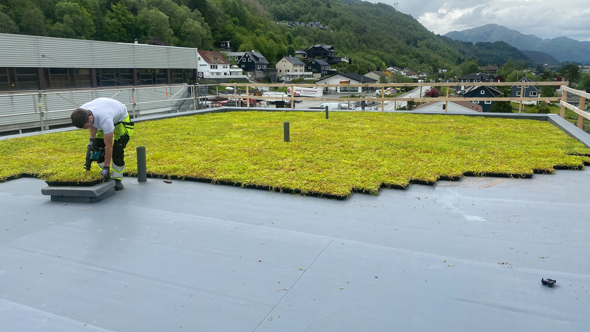 Montaż zielonego dachu z użyciem wkrętarki na płaskiej powierzchni. W tle zabudowa miejska i zalesione wzgórza.