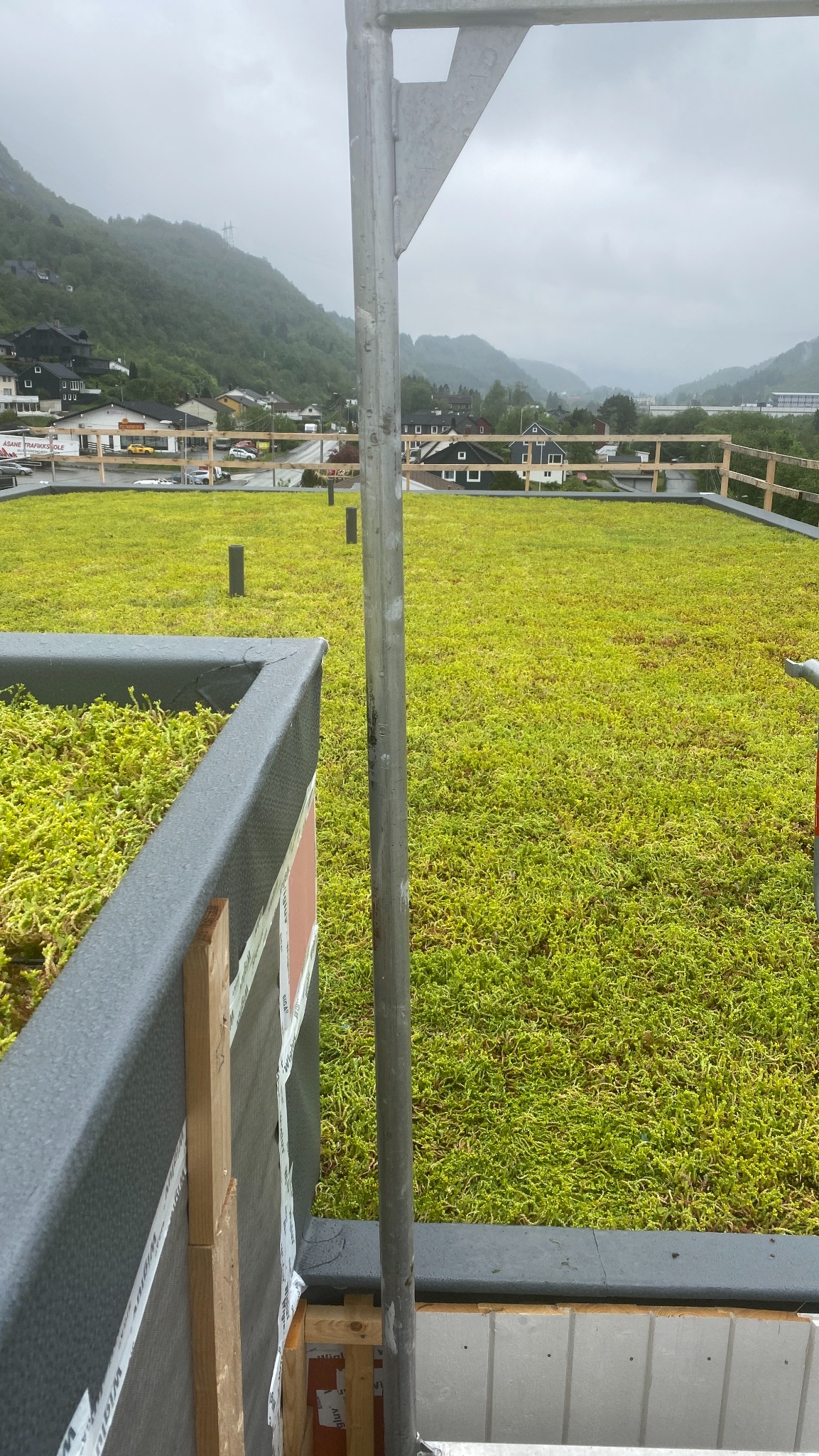 Zielony dach z roślinnością na tle gór i zabudowań, widok z góry, fragment rusztowania w kadrze. Ekologiczne rozwiązanie dachowe w otoczeniu natury.