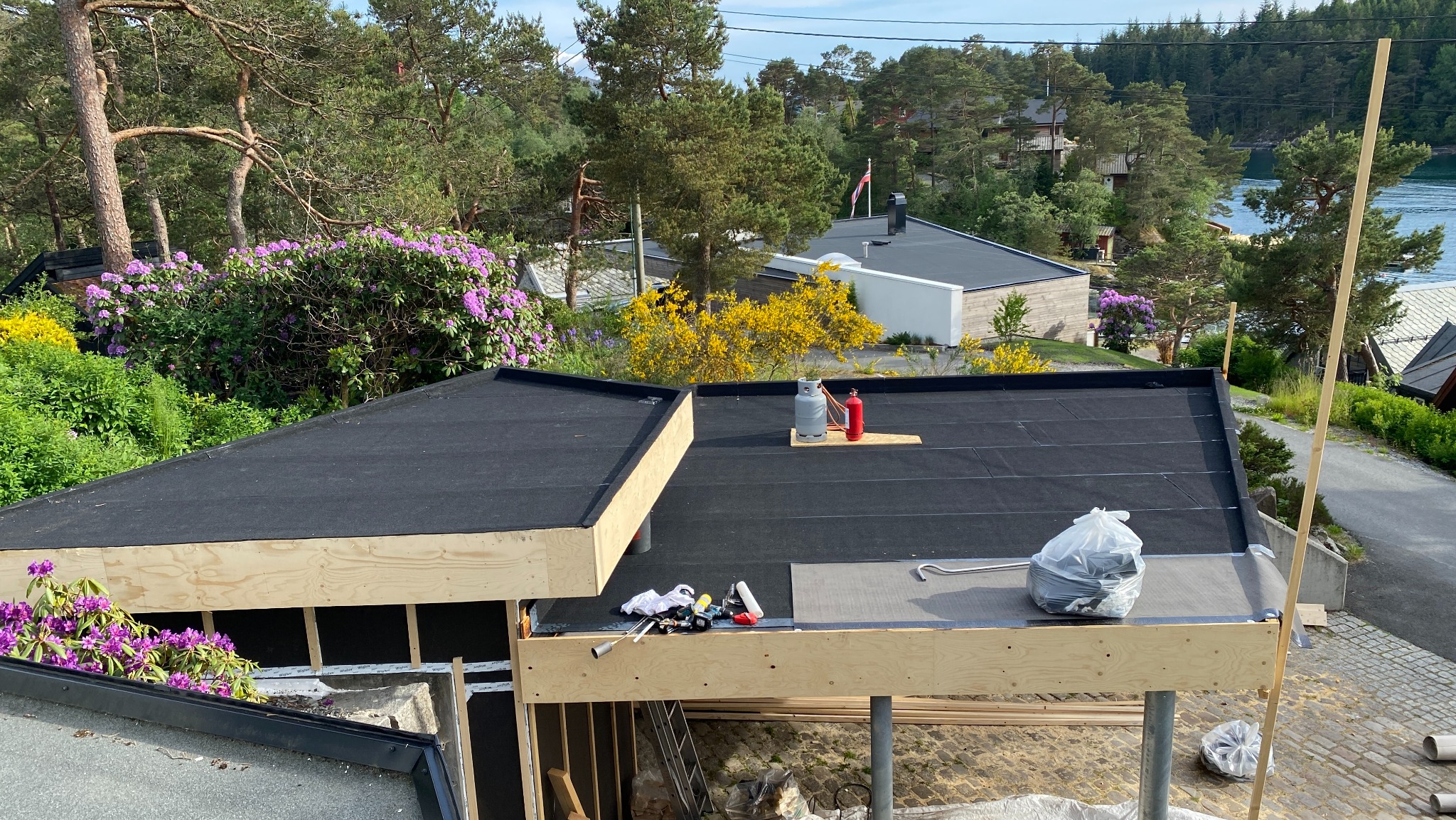 Czarny, płaski dach z widocznymi narzędziami i butlami gazowymi na tle zieleni i zbiornika wodnego. Dach w trakcie prac wykończeniowych, z elementami drewnianej konstrukcji.
