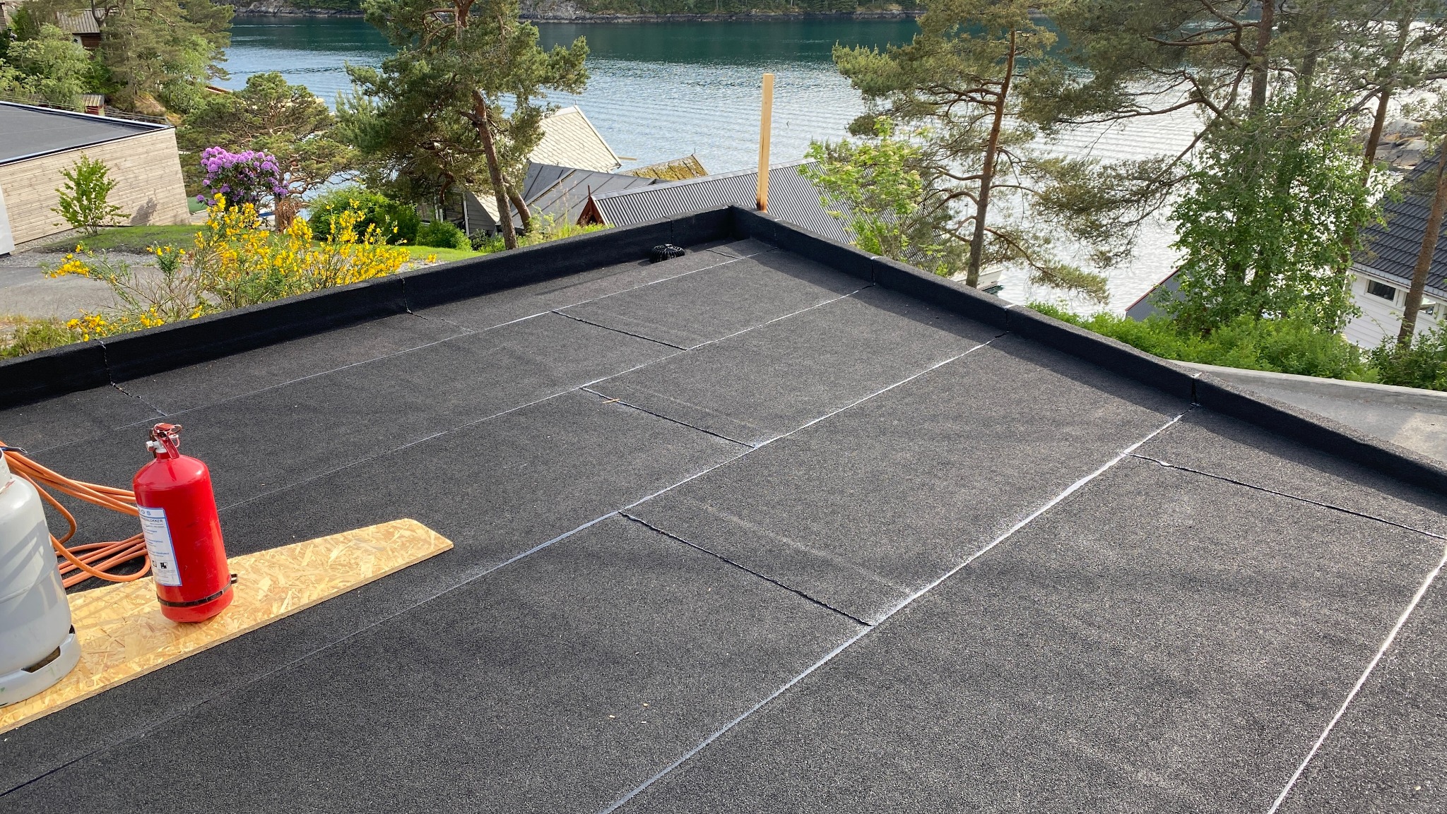Czarny, płaski dach pokryty papą termozgrzewalną z widocznymi łączeniami, butla gazowa i czerwona gaśnica na tle jeziora i zieleni.