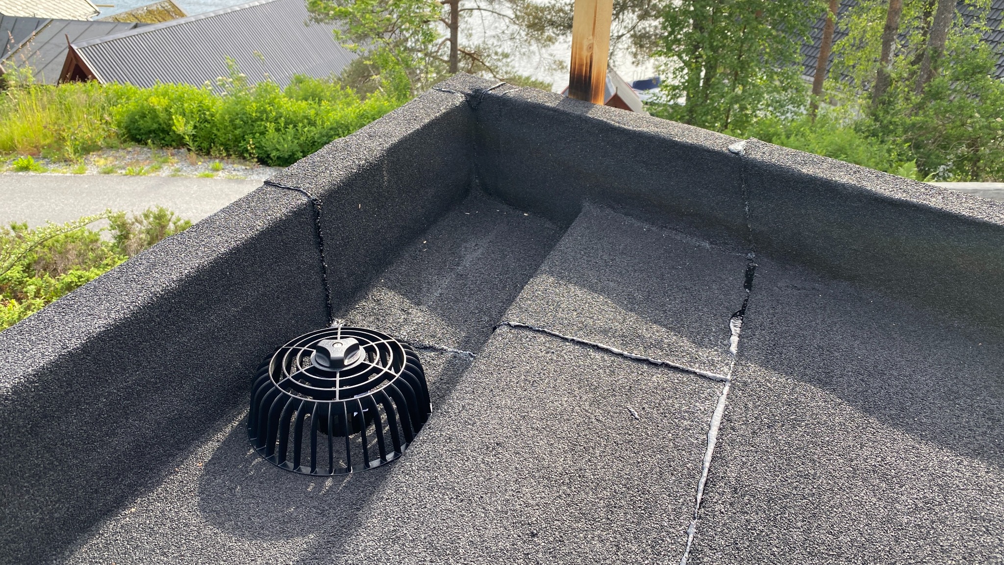 Czarny, płaski dach pokryty papą z wpustem dachowym. Widoczne łączenia papy i fragment otoczenia z zielenią w tle.