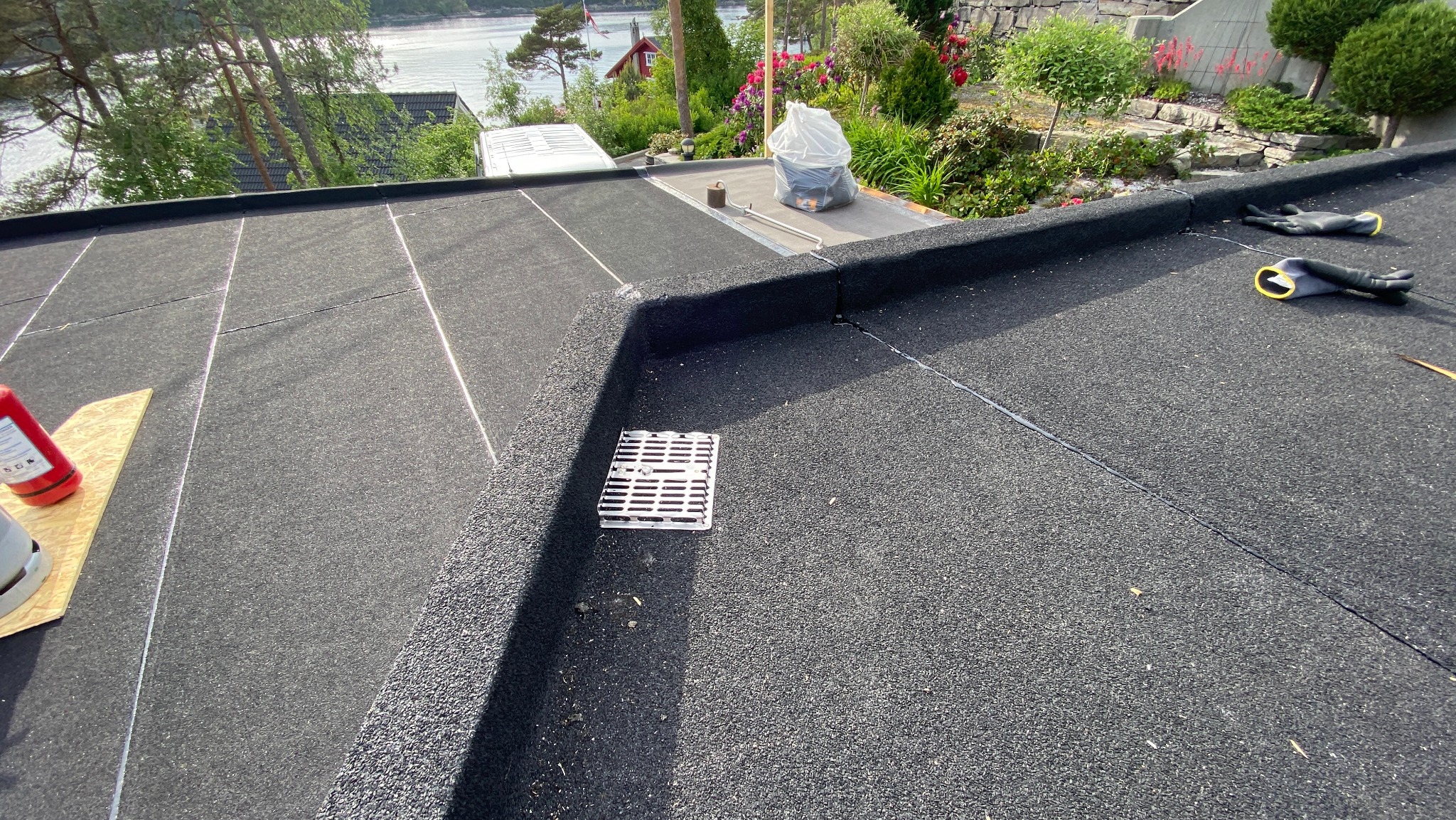 Czarny, płaski dach pokryty papą z widocznym odpływem, narzędziami i rękawicami roboczymi. W tle zieleń drzew i krzewów, co sugeruje otoczenie domu.