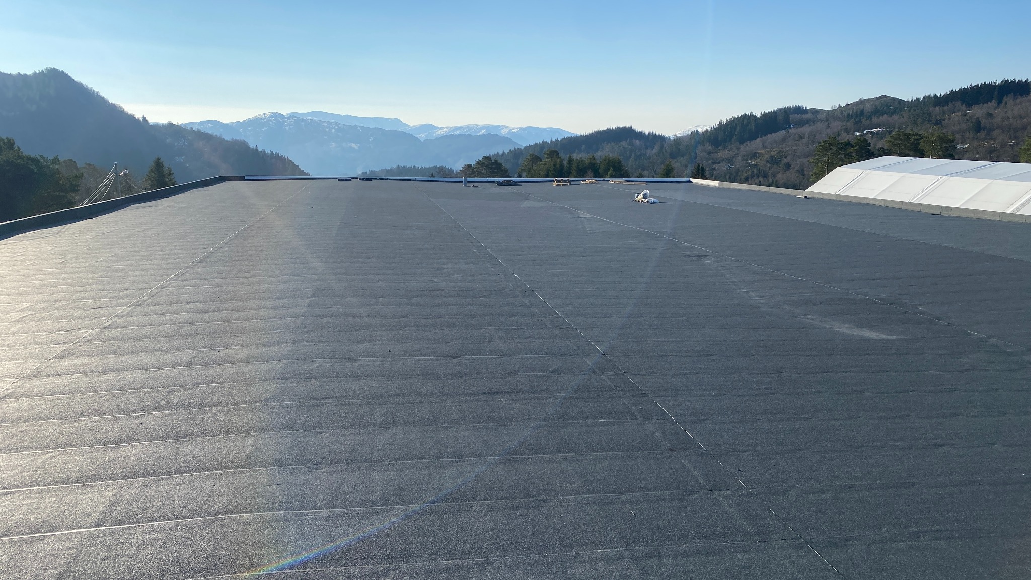 Czarny, płaski dach pokryty papą na tle gór i błękitnego nieba. Widoczne łączenia papy. Daleki plan ukazuje zalesione wzgórza i ośnieżone szczyty.