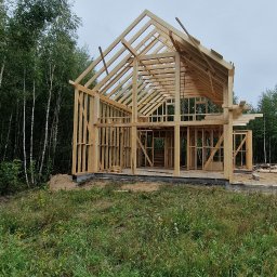 Z.p.u.h Tymek - Surowy drewniany szkielet domu jednorodzinnego w trakcie budowy, z widocznymi belkami konstrukcyjnymi i zarysami okien, otoczony zieloną trawą i drzewami.