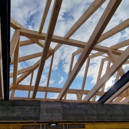 Z.p.u.h Tymek - Konstrukcja drewnianego dachu z widocznymi elementami stalowymi, na tle błękitnego nieba z chmurami, widok z wnętrza budynku w trakcie budowy.