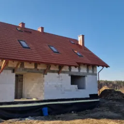 Surowy dom murowany z czerwoną dachówką, widoczne bloczki H+H oraz folia fundamentowa, w tle drugi dom i błękitne niebo.