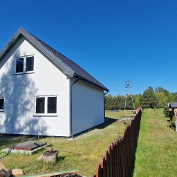 Z.p.u.h Tymek - Nowoczesny, biały dom z ciemnym dachem w słoneczny dzień. Widoczne drewniane elementy budowlane i narzędzia, sugerujące trwające prace wykończeniowe.