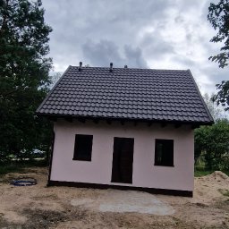 Z.p.u.h Tymek - Niewielki, parterowy budynek z ciemną dachówką, jasną elewacją i ciemnymi oknami, otoczony drzewami i piaskiem. Widok z dołu, pochmurne niebo.