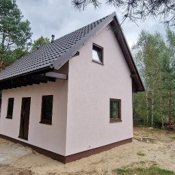 Z.p.u.h Tymek - Niewielki, otynkowany dom z ciemną dachówką i brązowymi oknami, otoczony zielenią drzew. Widoczne elementy wykończeniowe i fragment terenu.