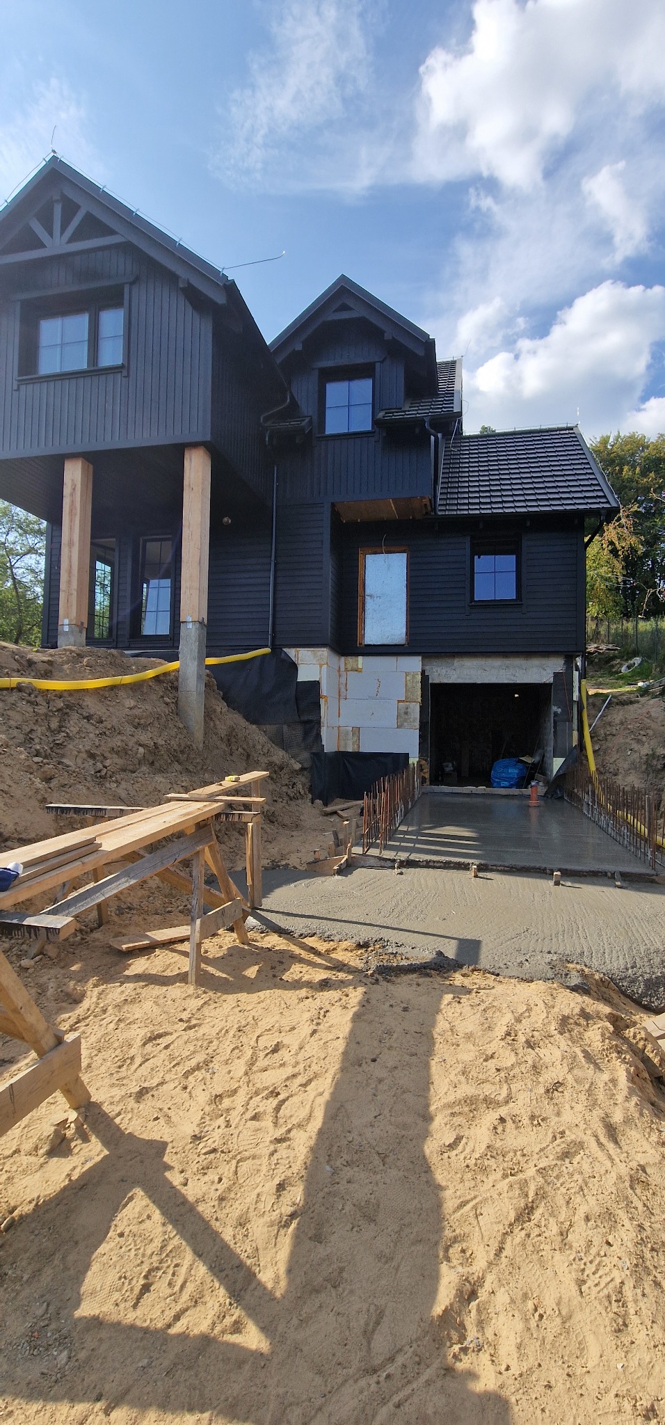 Dwukondygnacyjny, ciemny dom w stylu skandynawskim w trakcie budowy, z widocznym wjazdem do garażu, świeżo wylanym betonem, częściowo ocieplonym fundamentem i żółtą rurą osłonową wzdłuż ściany.