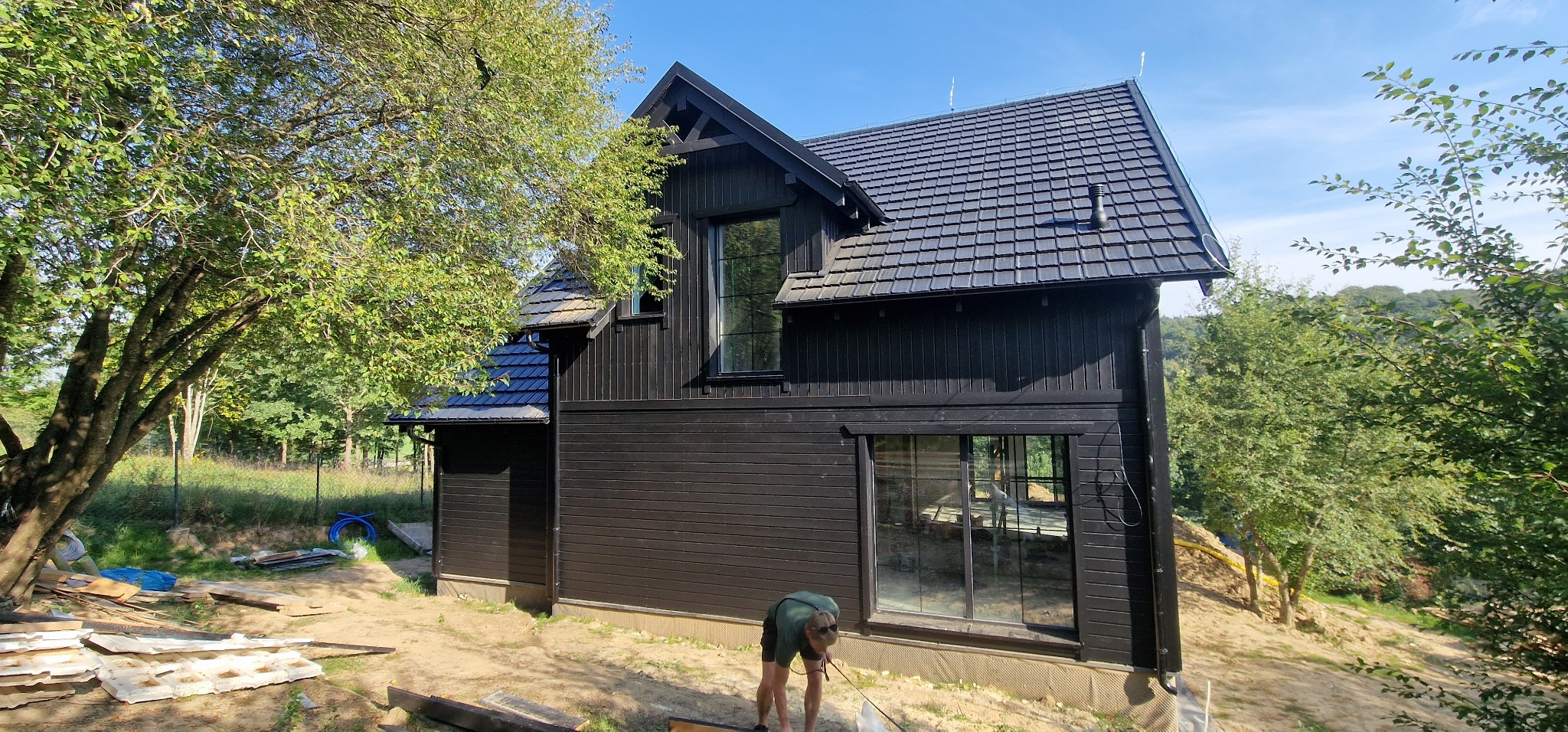 Czarny, drewniany dom z ciemną dachówką w trakcie budowy, widoczny pracownik przy ziemi i porozrzucane materiały budowlane.