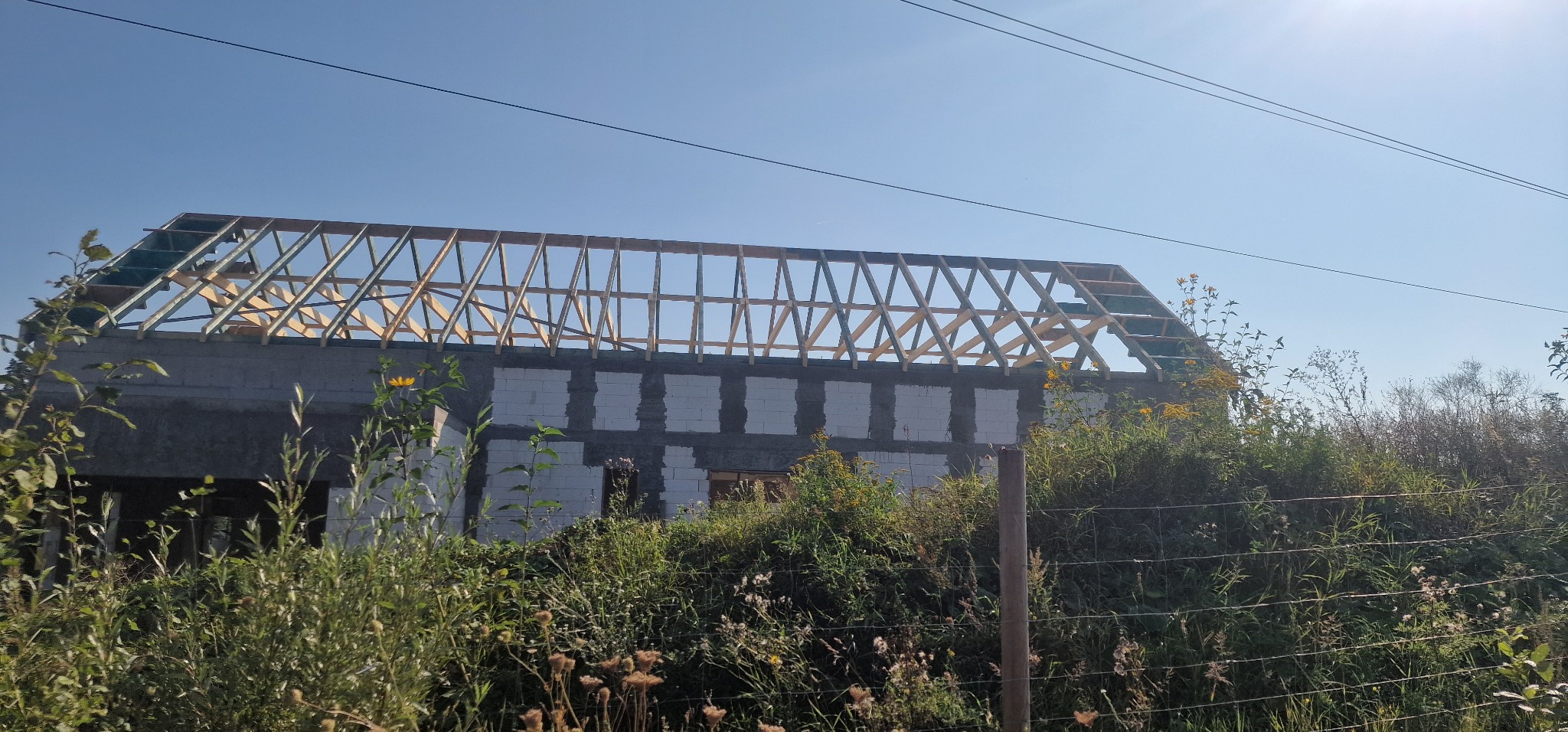 Niedokończony budynek z białych bloczków z drewnianą konstrukcją dachu, widoczny przez bujną roślinność i ogrodzenie z siatki.