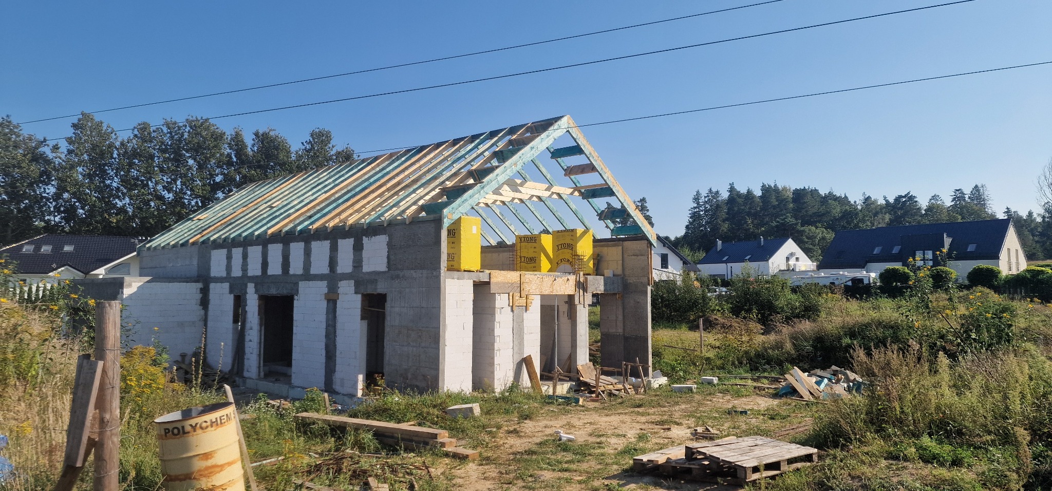 Wznoszenie domu murowanego z bloczków YTONG, widoczna konstrukcja dachu z deskowaniem, żółte paczki materiału izolacyjnego, w tle inne budynki i drzewa.