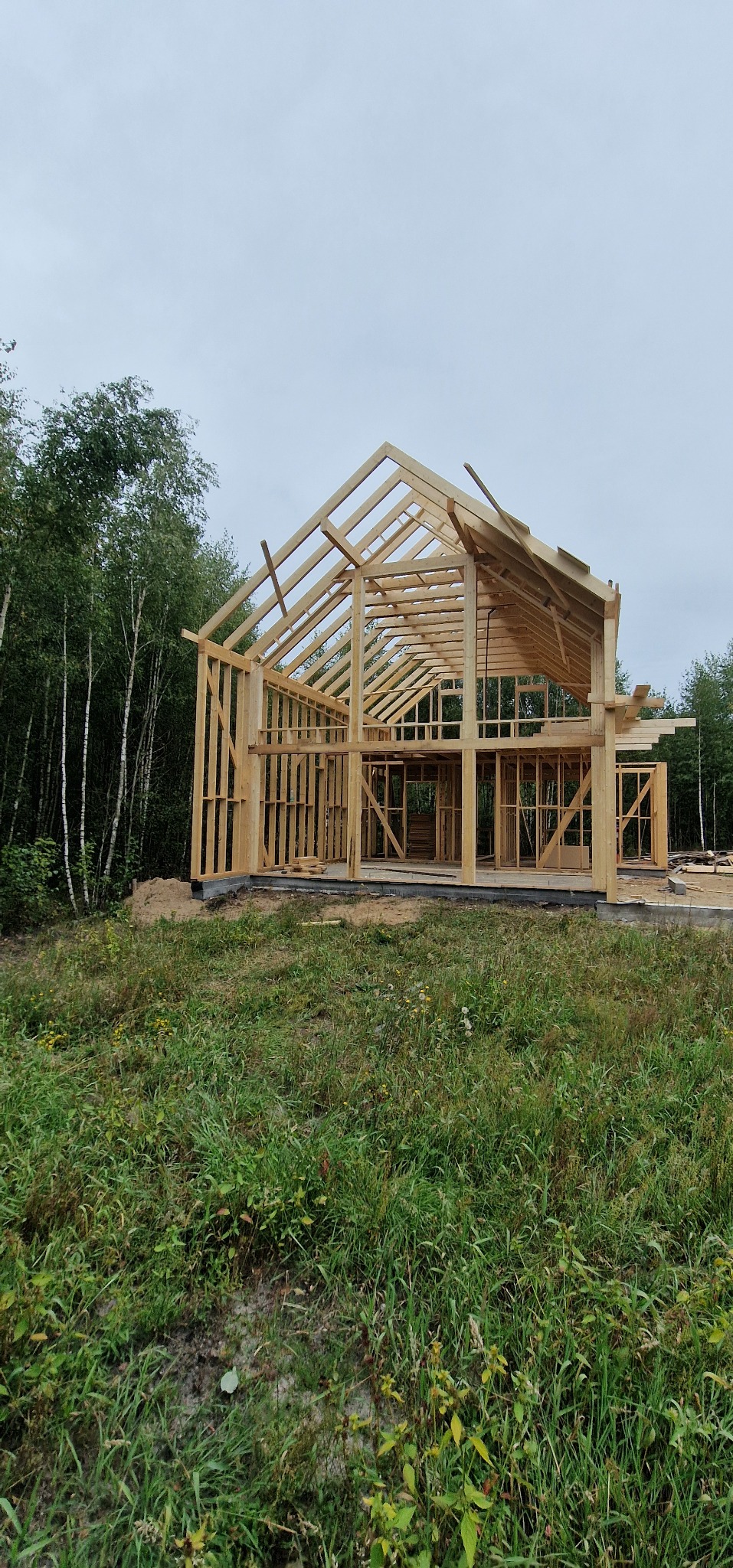 Surowy drewniany szkielet domu jednorodzinnego w trakcie budowy, z widocznymi belkami konstrukcyjnymi i zarysami okien, otoczony zieloną trawą i drzewami.