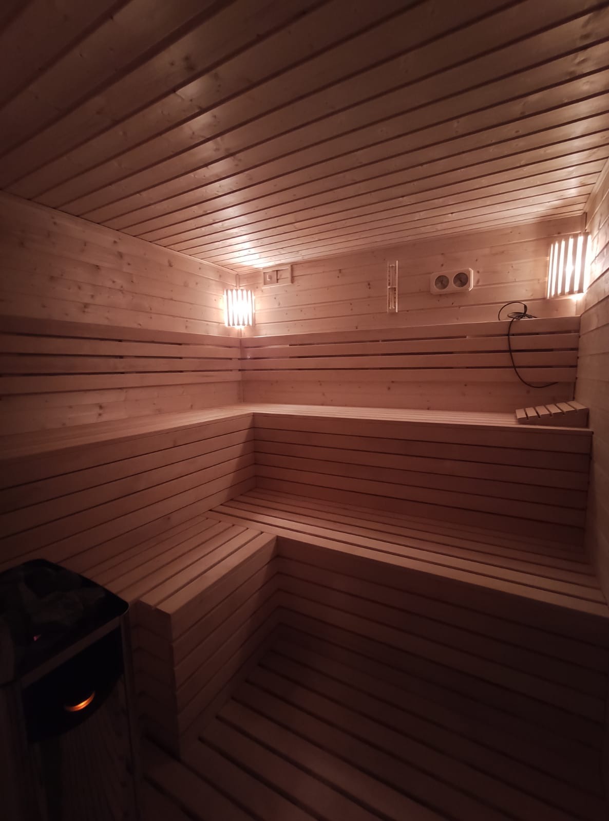 Wnętrze sauny wykończonej drewnem w Dąbrowie, z piecem elektrycznym w lewym dolnym rogu i oświetleniem ściennym.