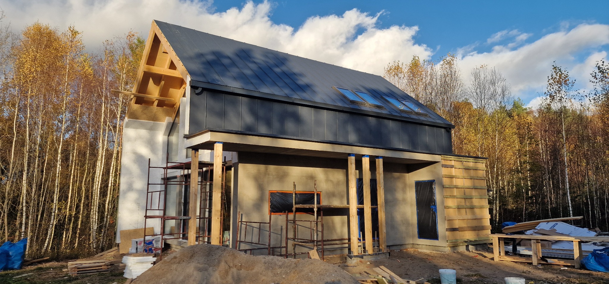 Dom w trakcie budowy: widoczne ściany z betonu komórkowego, drewniana konstrukcja dachu bez pokrycia, część elewacji obłożona drewnem, rusztowania i materiały budowlane na placu budowy, jesienne...