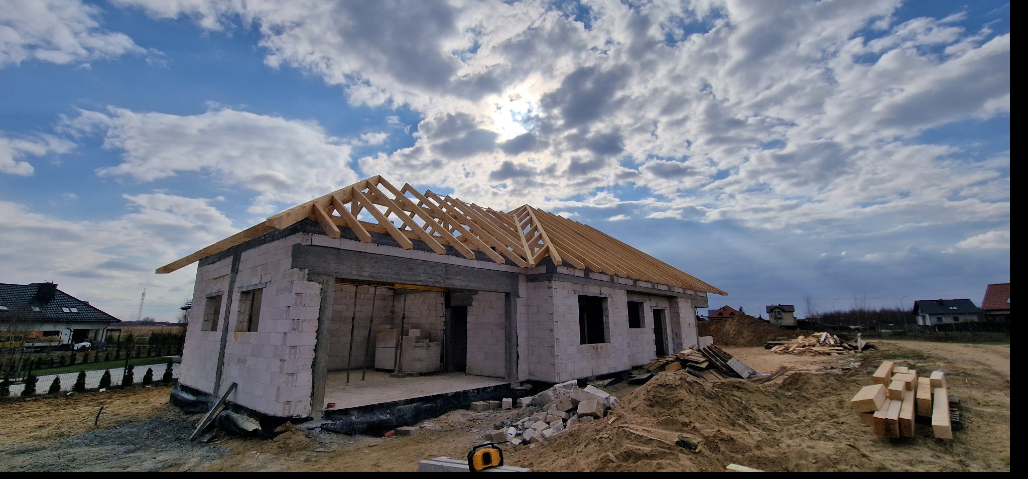 Budowa domu murowanego w trakcie realizacji: widoczne ściany z bloczków, drewniana konstrukcja dachu w trakcie montażu oraz stos drewna na placu budowy, niebo z chmurami w tle.