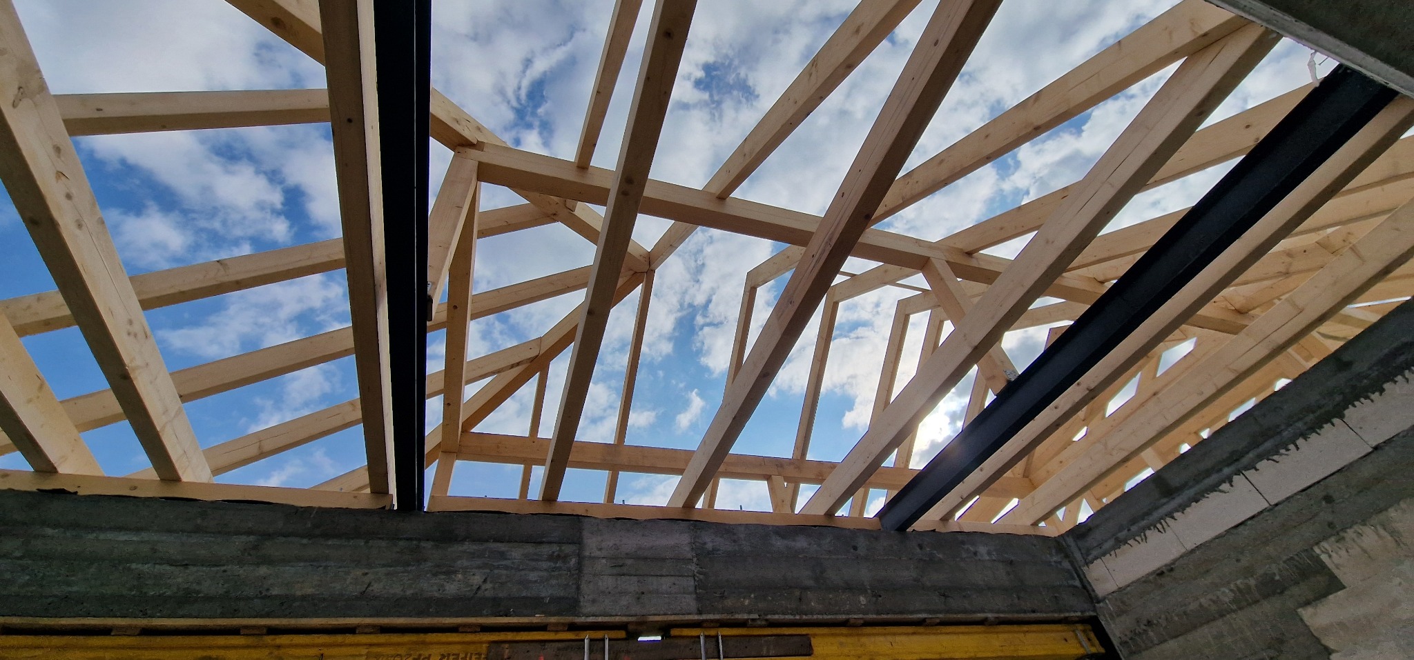 Konstrukcja drewnianego dachu z widocznymi elementami stalowymi, na tle błękitnego nieba z chmurami, widok z wnętrza budynku w trakcie budowy.