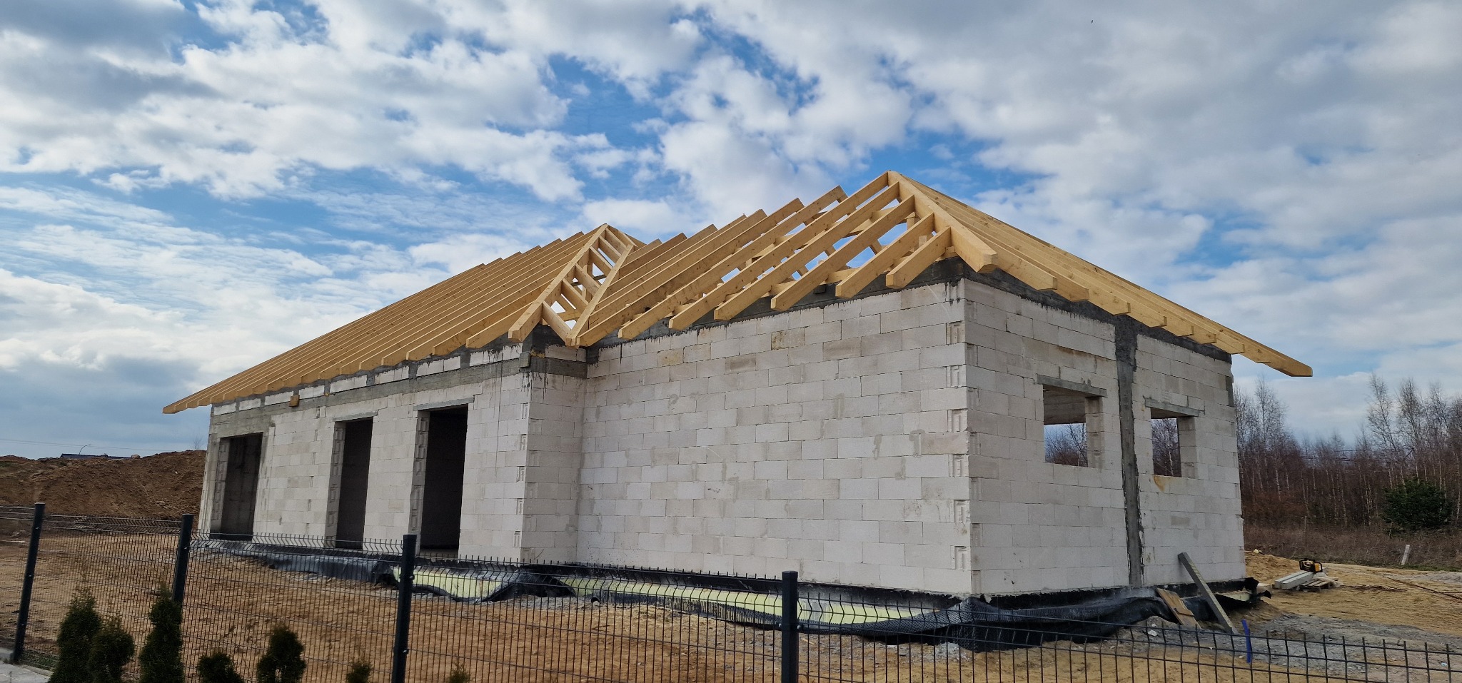 Surowy stan domu z białych bloczków z widoczną konstrukcją drewnianą dachu, otwartymi otworami okiennymi i drzwiowymi, ogrodzony siatką.