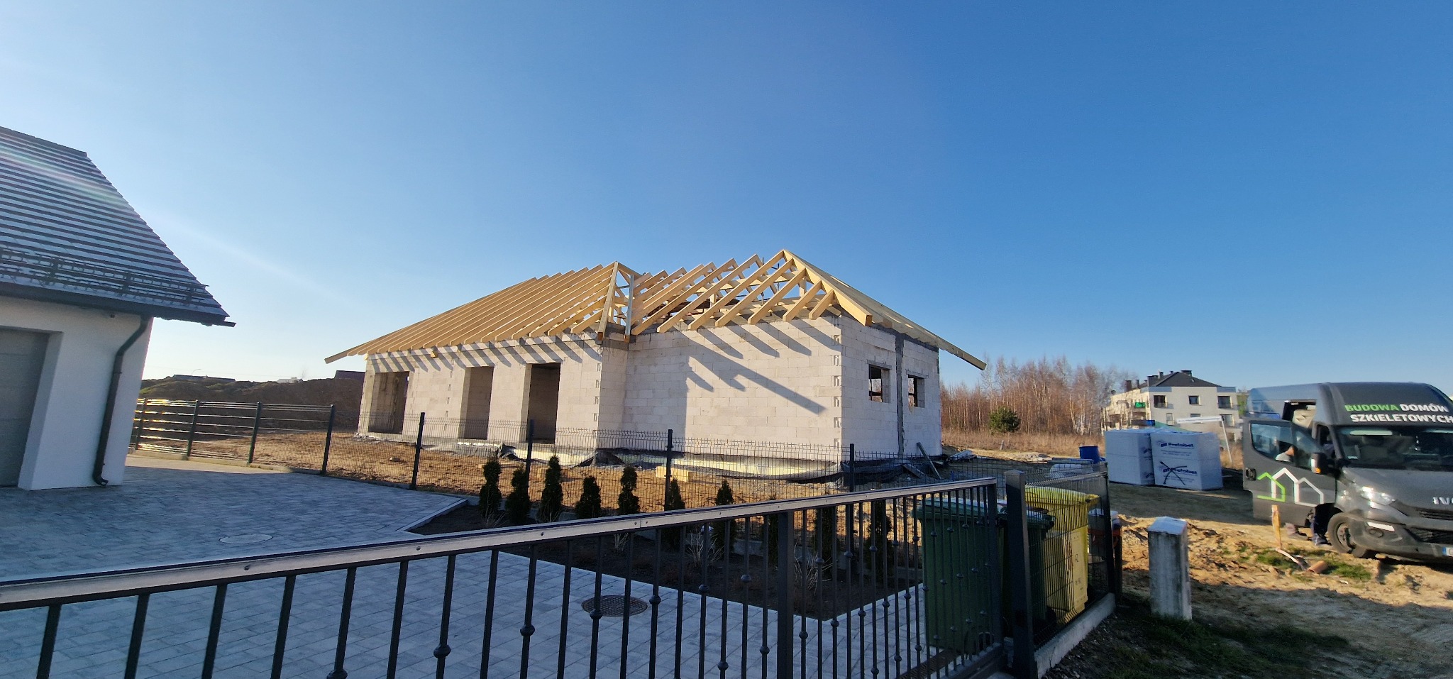 Wznoszony dom z białego bloczka z widoczną konstrukcją dachu z drewnianych krokwi, ogrodzony siatką, na pierwszym planie fragment metalowego ogrodzenia i ozdobne tuje, w tle zaparkowany bus...