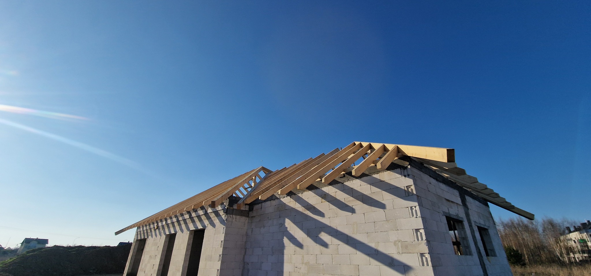 Wznoszenie drewnianej konstrukcji dachu na domu z białych bloczków, widoczne otwory okienne, błękitne niebo w tle.