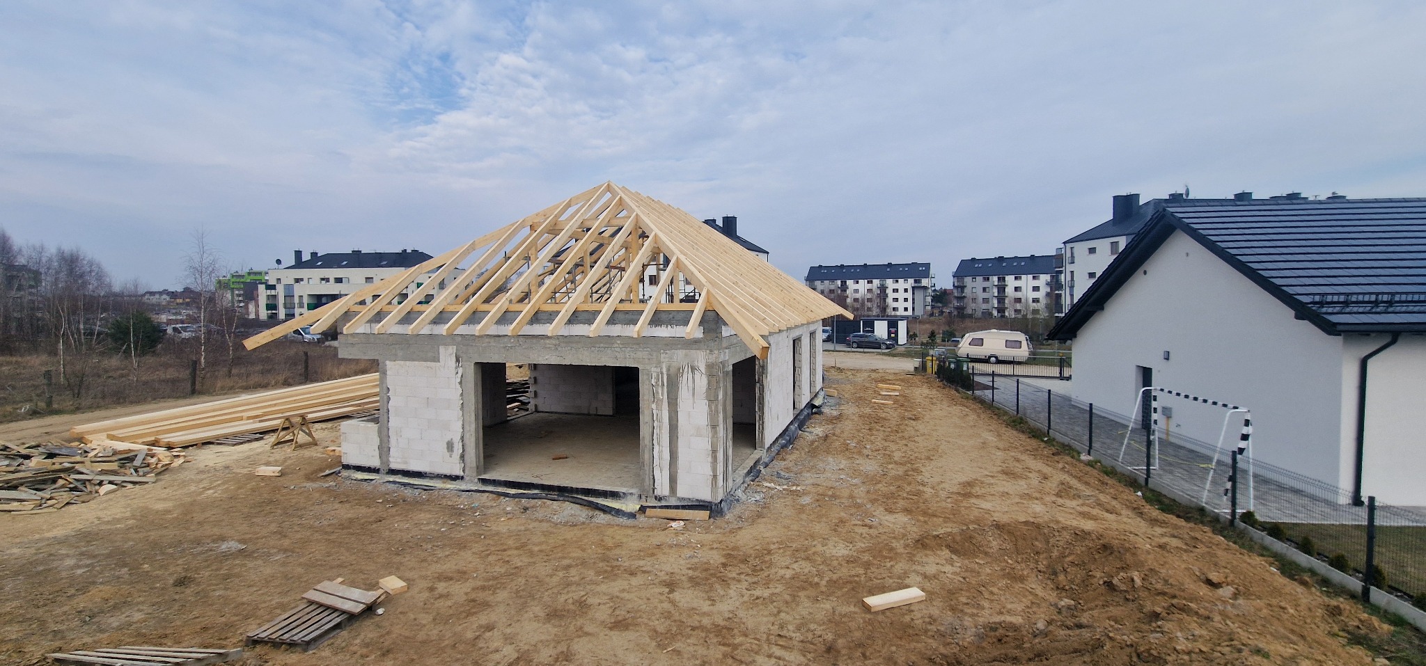 Surowy stan domu z betonu komórkowego z drewnianą konstrukcją dachu, na tle zabudowy wielorodzinnej i błękitnego nieba.
