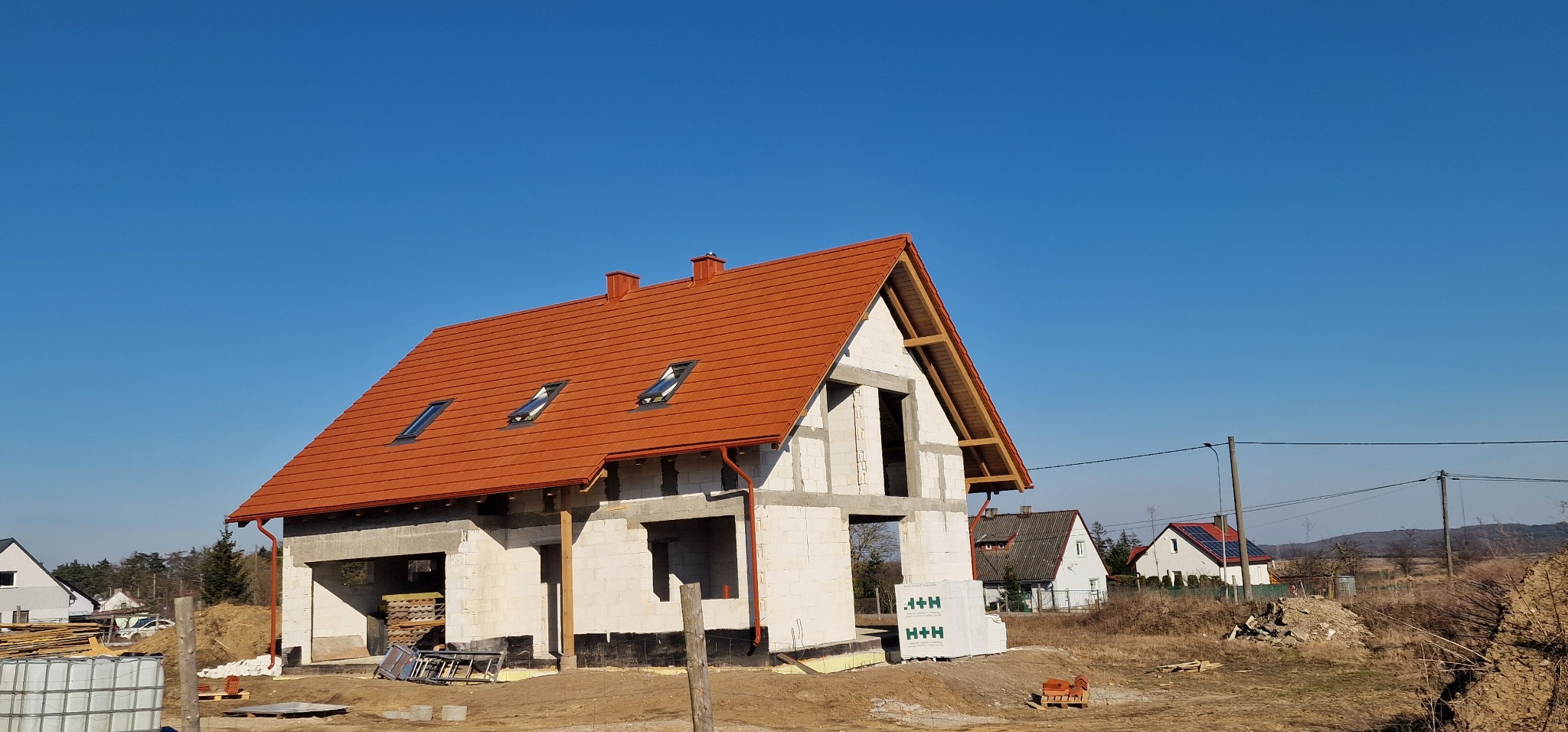 Budowa domu z białych bloczków, widoczny dach z czerwonej dachówki i okna dachowe, na pierwszym planie paleta z bloczkami H+H, w tle inne domy i błękitne niebo.
