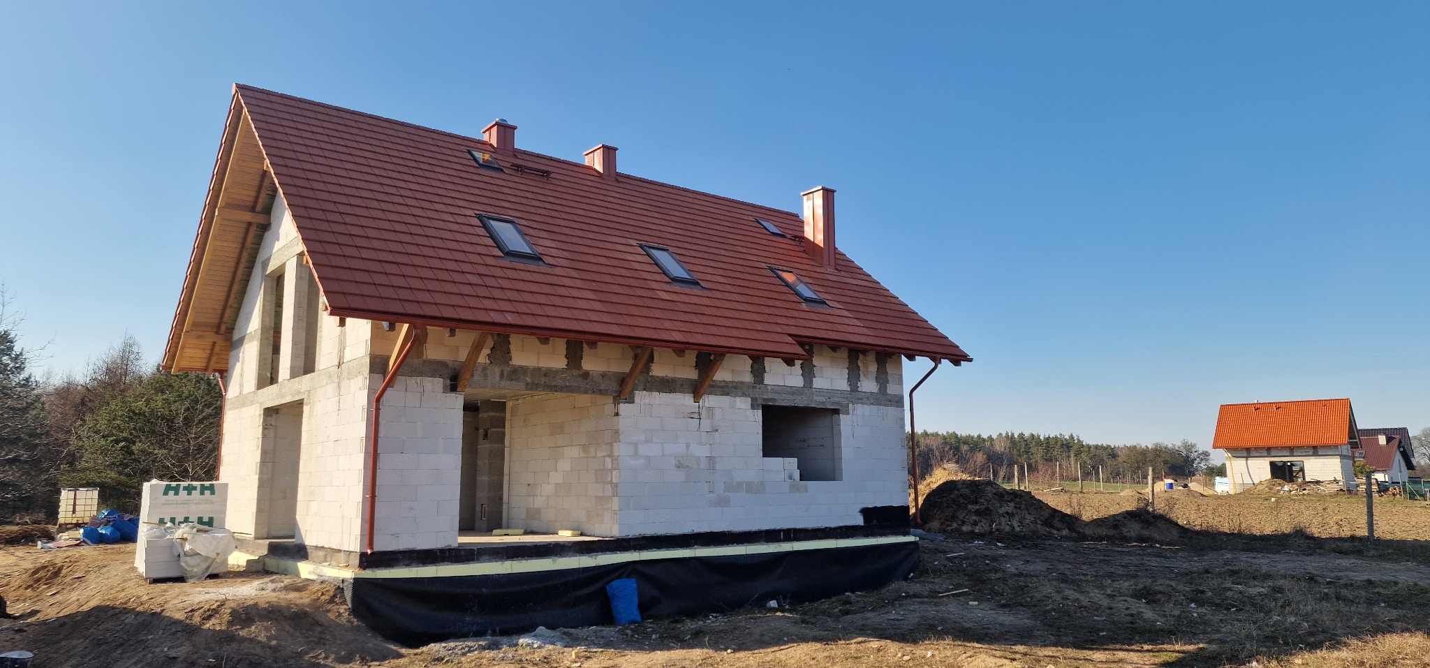 Surowy dom murowany z czerwoną dachówką, widoczne bloczki H+H oraz folia fundamentowa, w tle drugi dom i błękitne niebo.