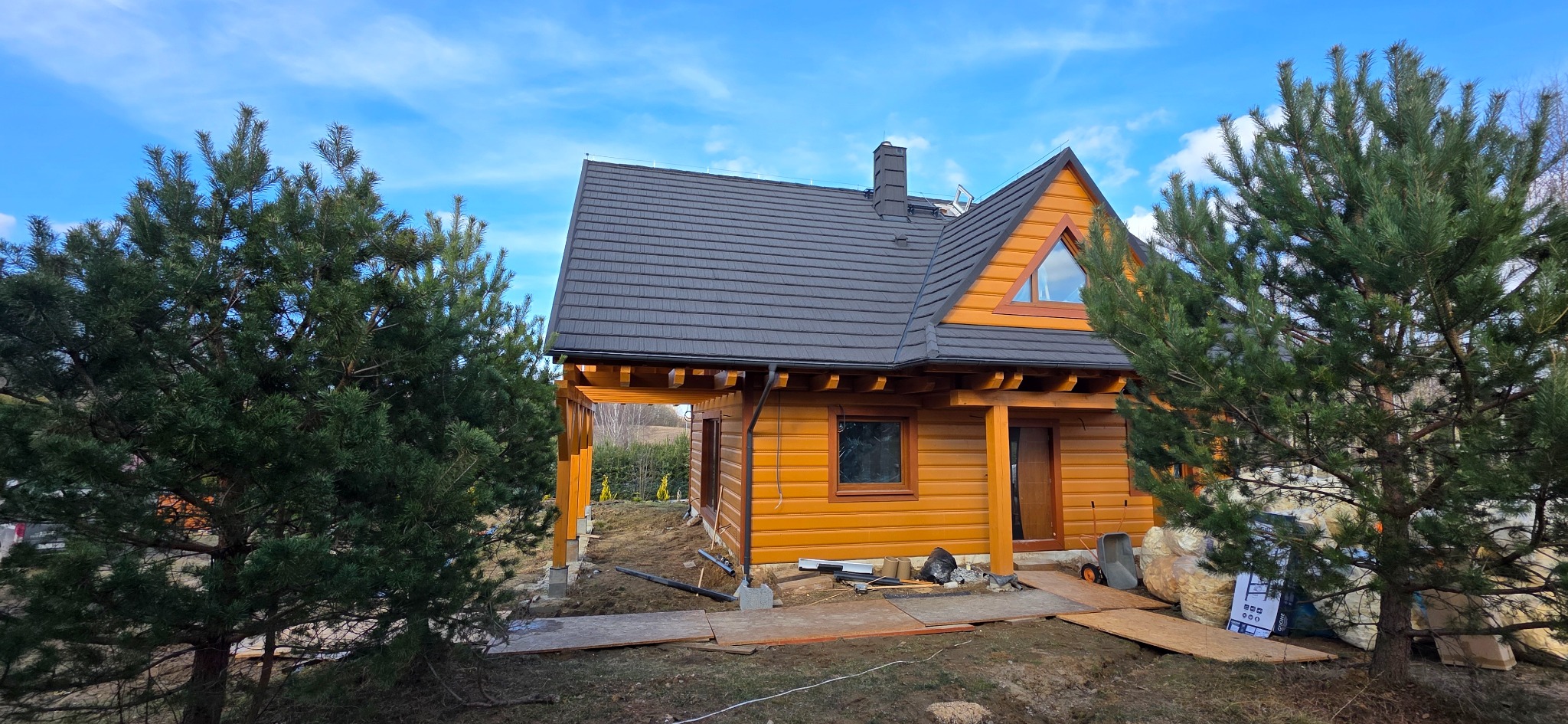 Drewniany dom z ciemnym dachem, widoczny z zewnątrz, otoczony zielenią drzew iglastych i elementami placu budowy. Uwagę przykuwa trójkątne okno na szczycie.