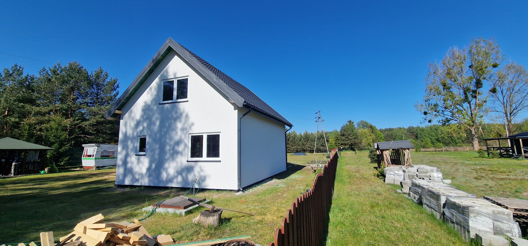 Nowoczesny, biały dom z ciemnym dachem w słoneczny dzień. Widoczne drewniane elementy budowlane i narzędzia, sugerujące trwające prace wykończeniowe.