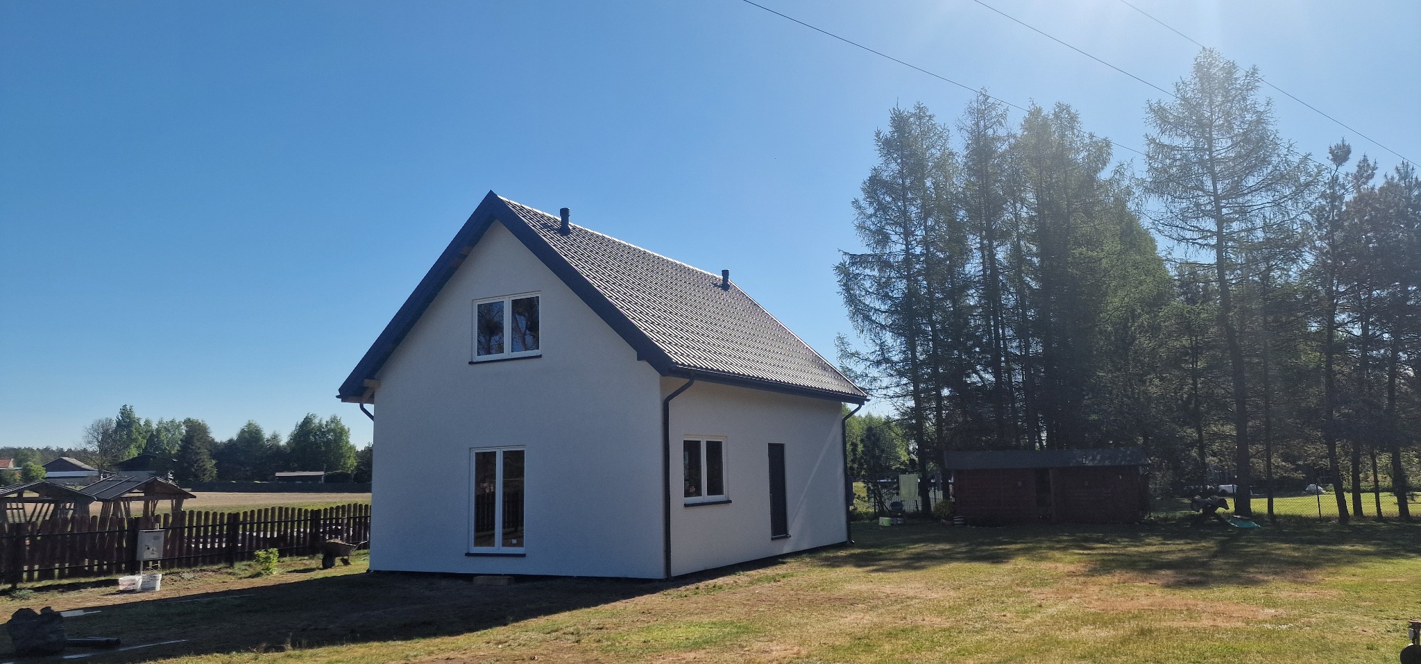 Biały, dwukondygnacyjny dom z ciemnym dachem na tle błękitnego nieba i zieleni drzew. Widoczne okna i rynny. Dom otoczony trawnikiem. Słoneczny dzień.