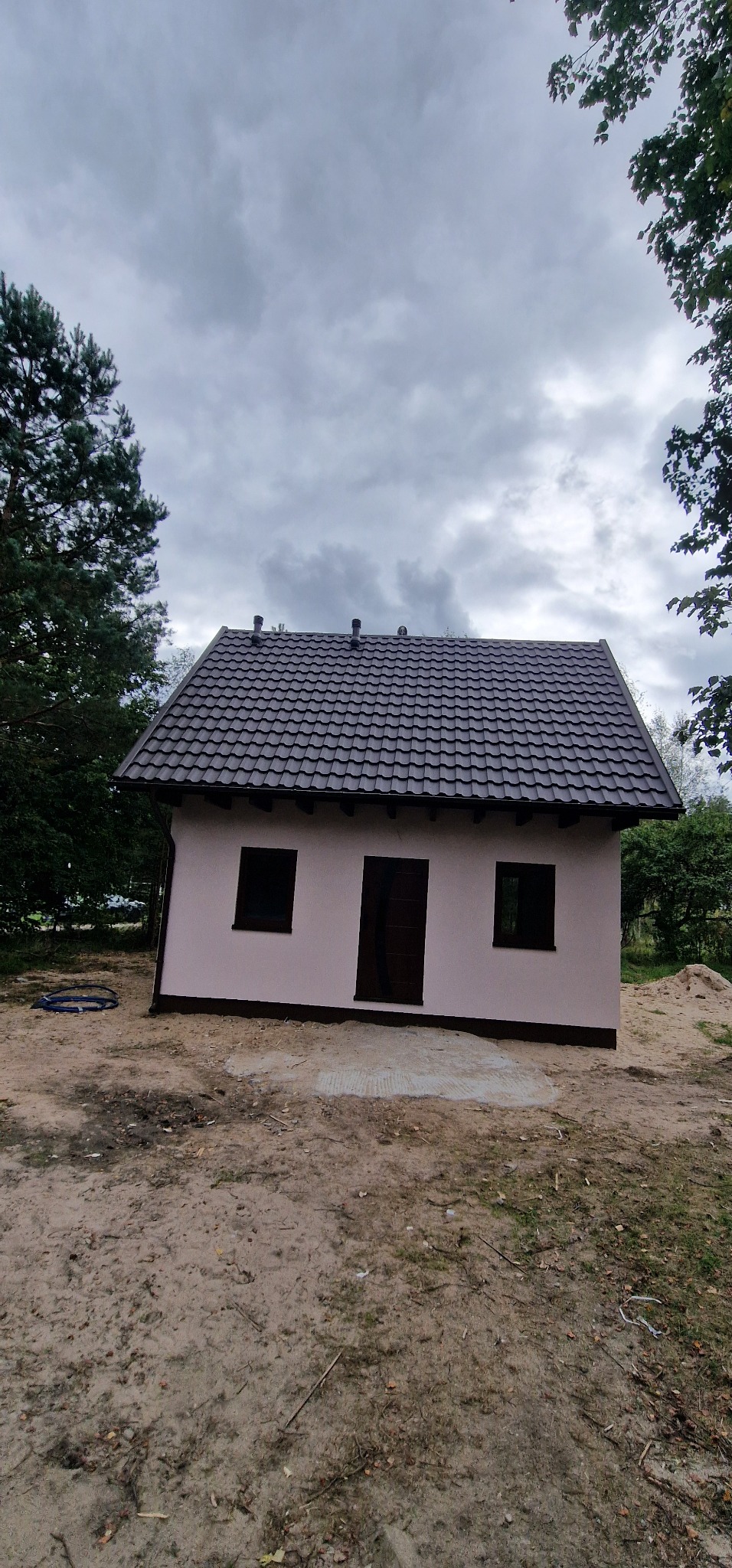 Niewielki, parterowy budynek z ciemną dachówką, jasną elewacją i ciemnymi oknami, otoczony drzewami i piaskiem. Widok z dołu, pochmurne niebo.