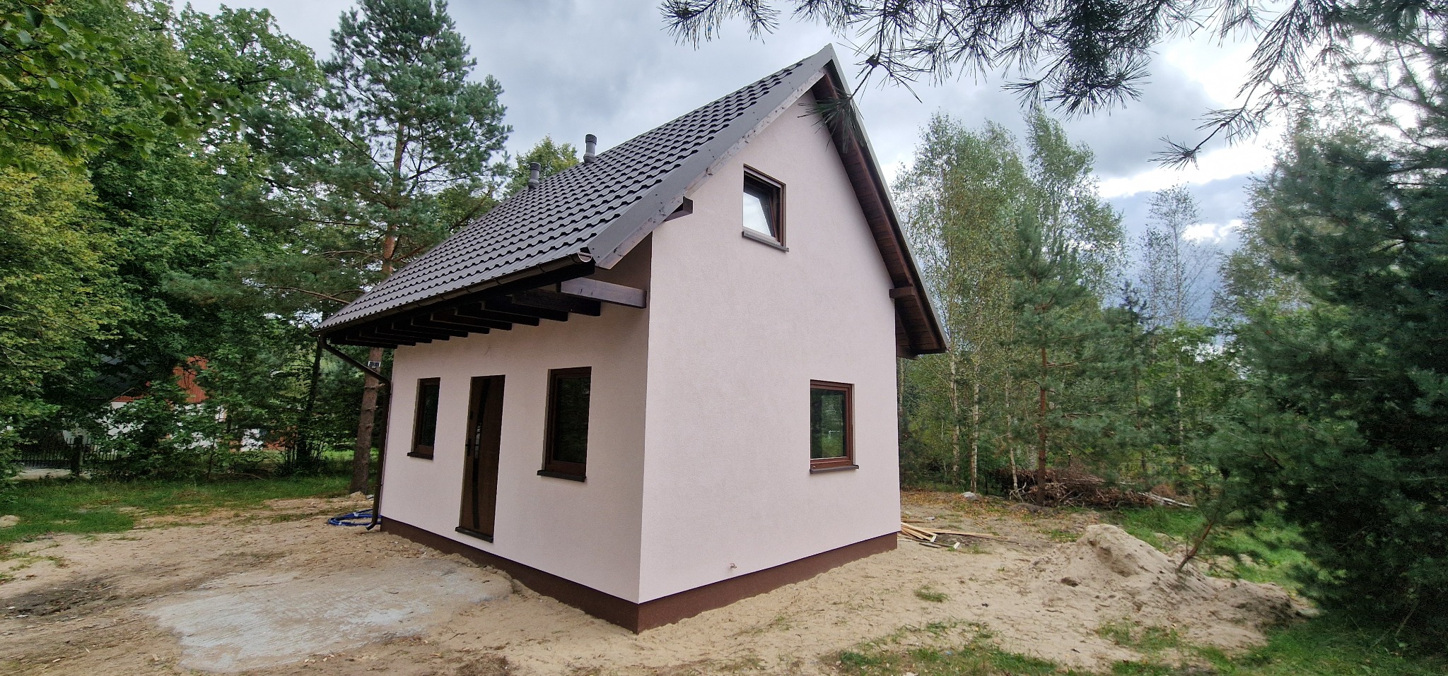 Niewielki, otynkowany dom z ciemną dachówką i brązowymi oknami, otoczony zielenią drzew. Widoczne elementy wykończeniowe i fragment terenu.
