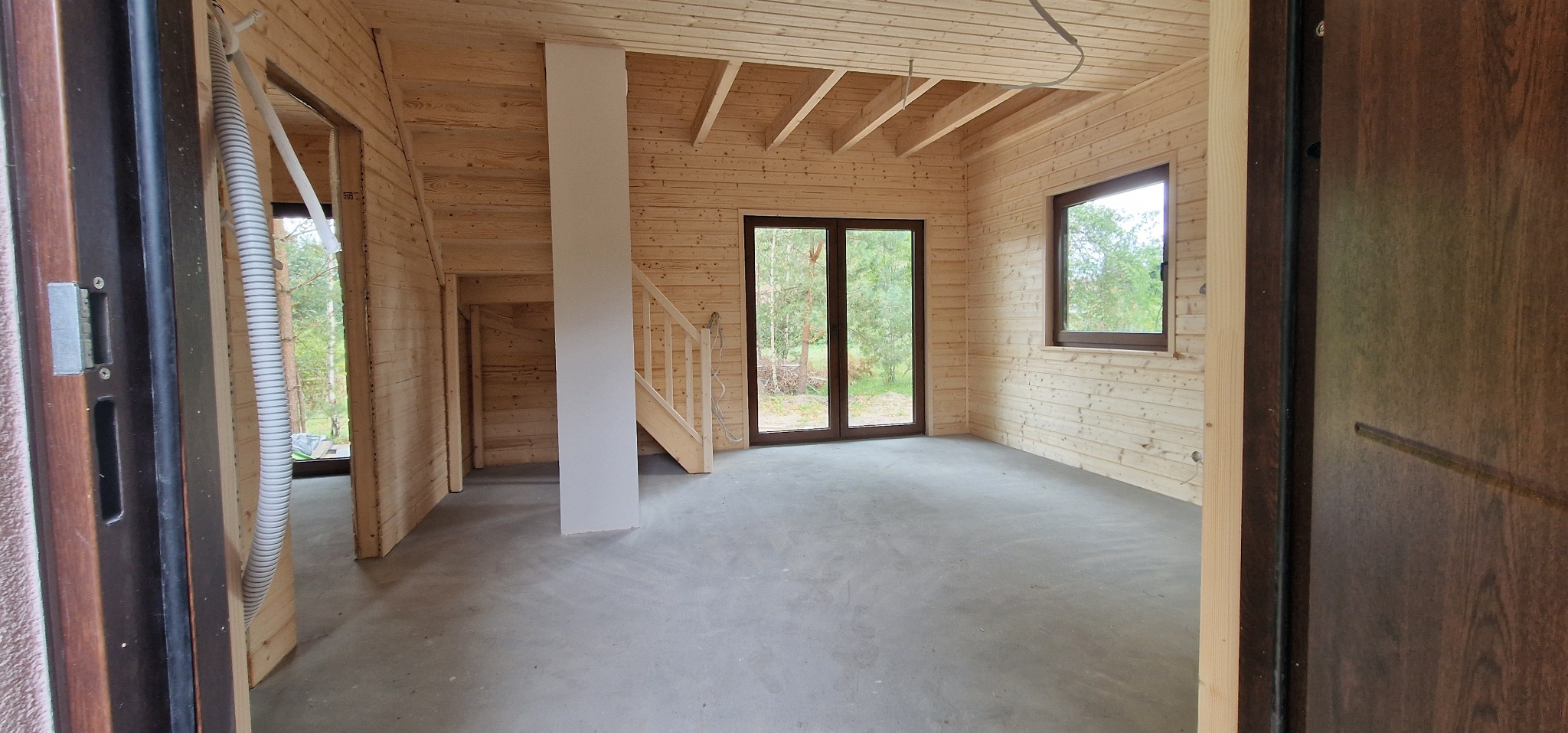 Wnętrze domu szkieletowego w trakcie budowy: surowe drewniane ściany, okna z widokiem na zieleń, betonowa podłoga, schody i pionowa kolumna. Widoczne elementy instalacji.