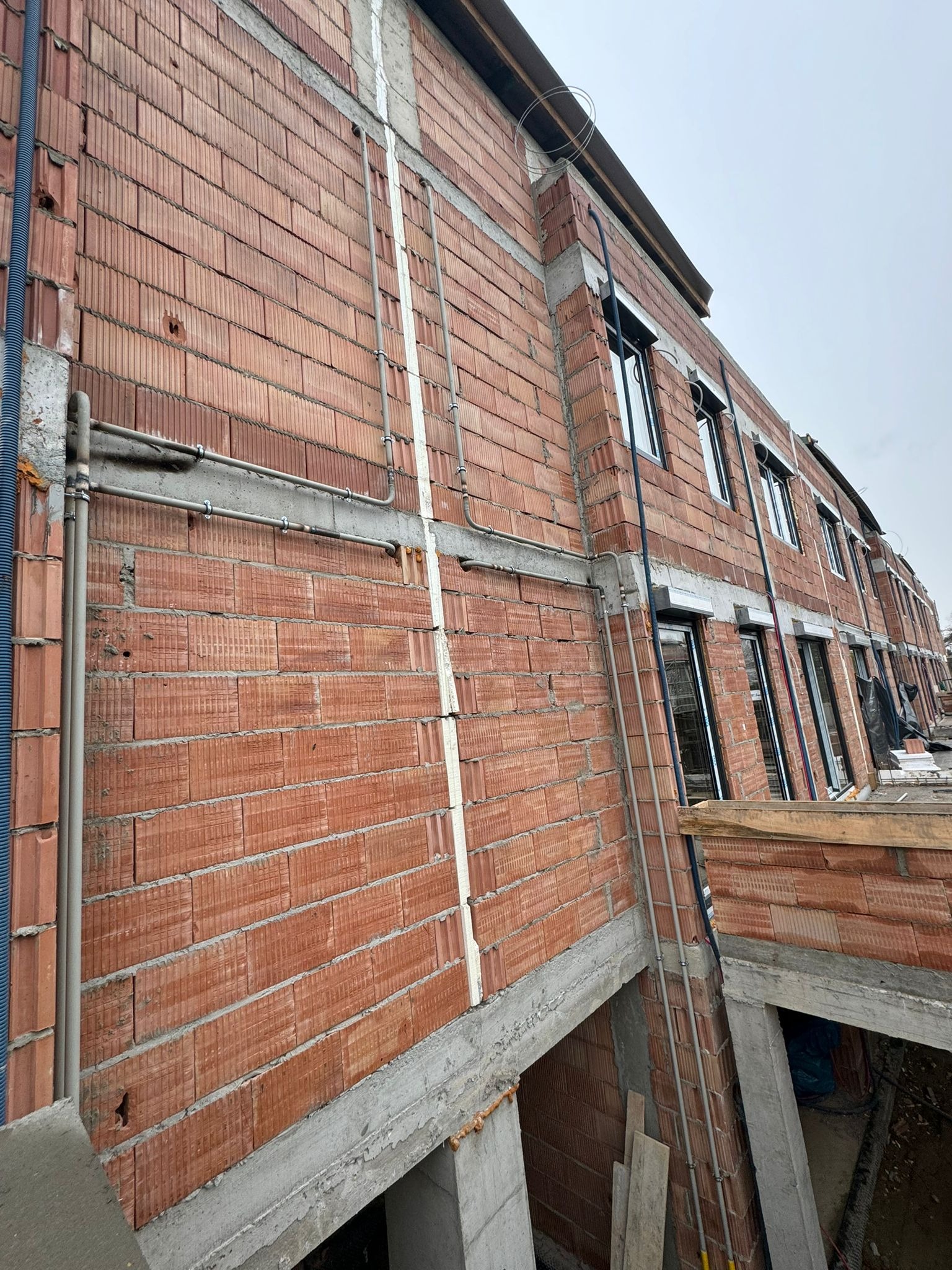 Surowa ściana z czerwonej cegły z widocznymi rurami instalacyjnymi prowadzonymi na zewnątrz budynku, okna w trakcie montażu, stan surowy.