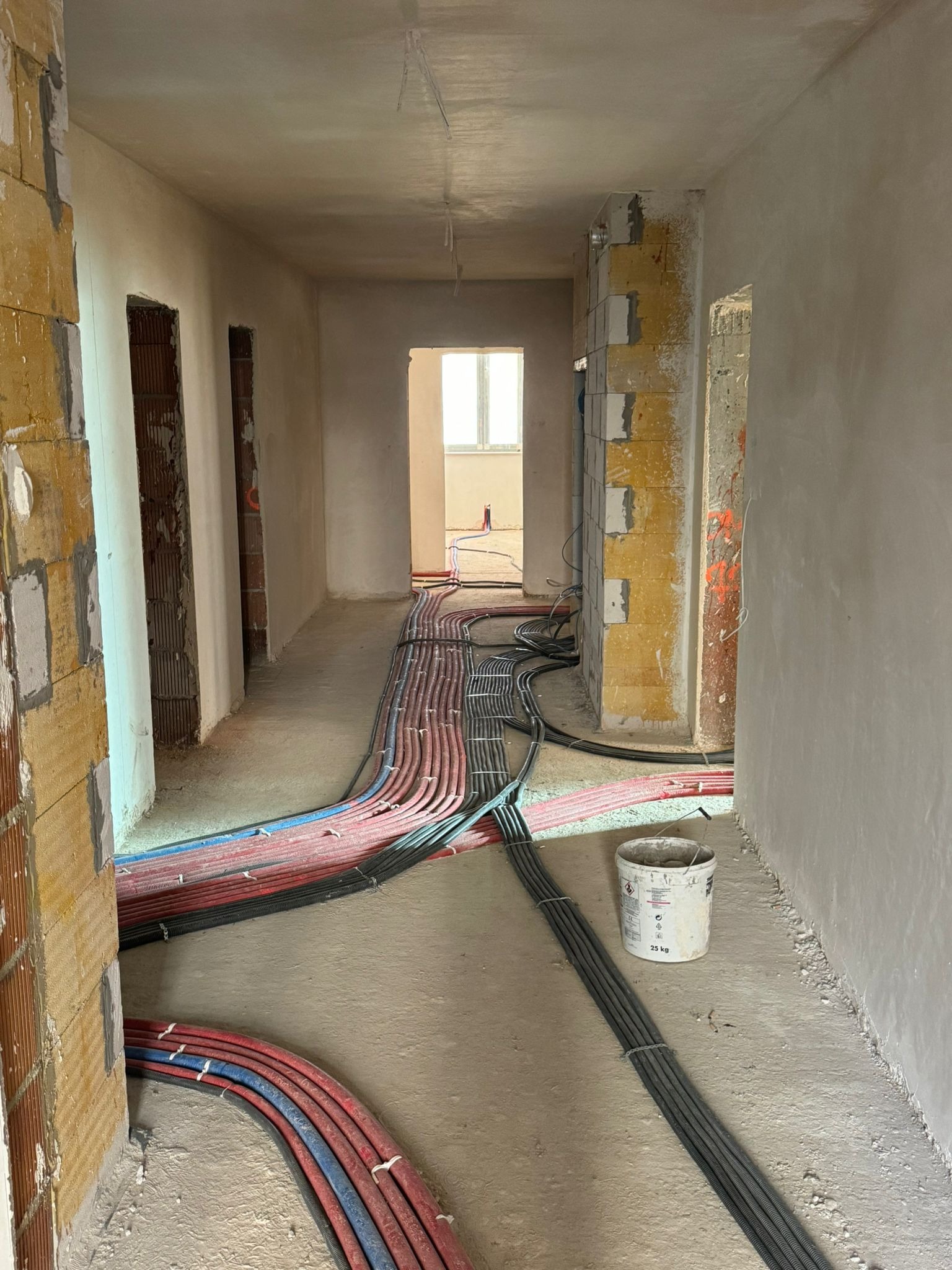 Wnętrze w trakcie prac instalacyjnych: wiązki rur w kolorach czerwonym, niebieskim i czarnym prowadzone wzdłuż korytarza, ściany w stanie surowym, widoczny pustak ceramiczny, wiadro z zaprawą.